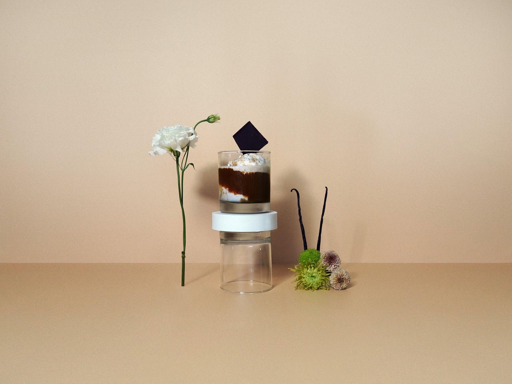A small, clear glass jar with a white lid containing fragrance oil. A flower and a vanilla bean are placed next to the jar.