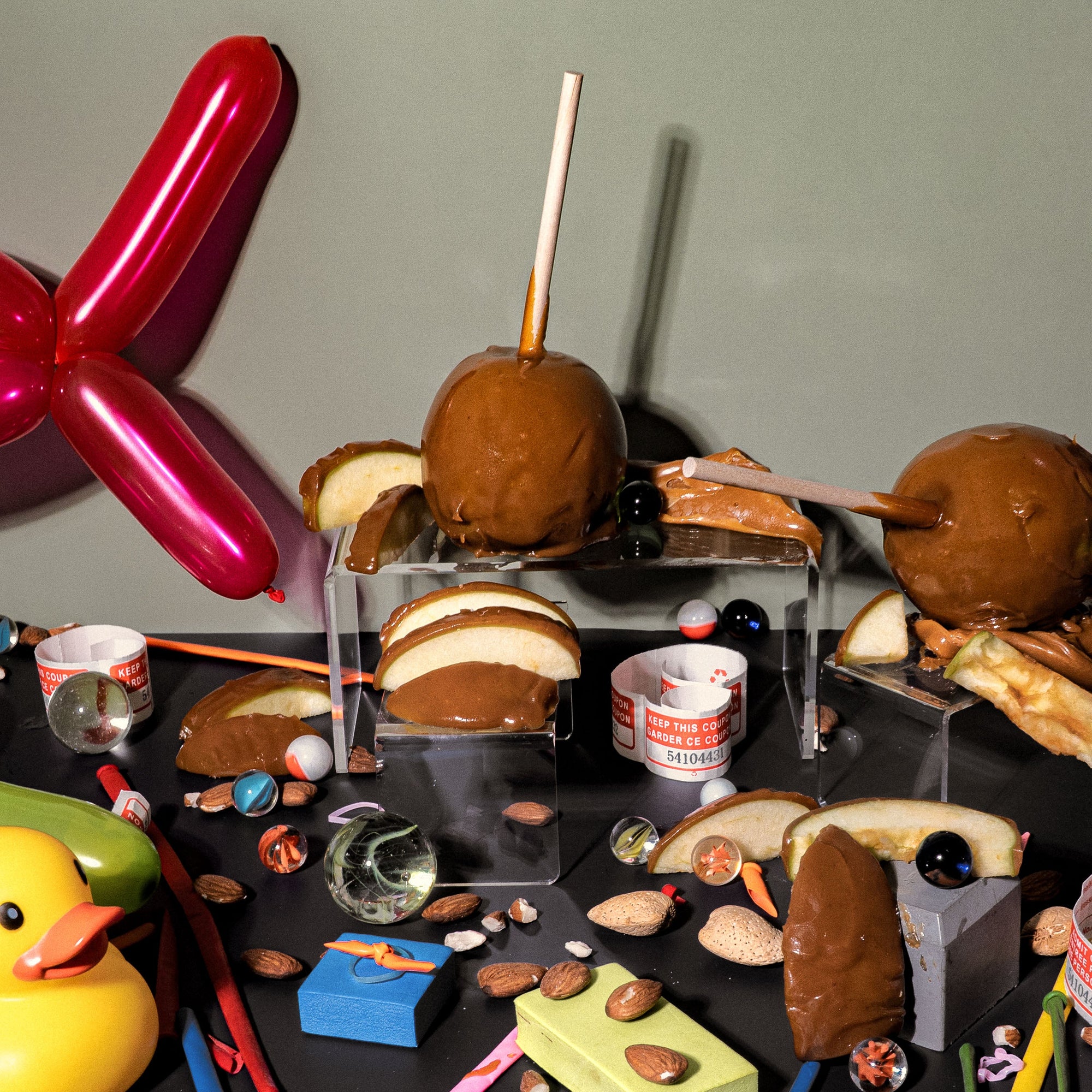 Whimsical flat lay, for a fragrance oil, featuring caramel apples, sliced green apples, marbles, almonds, carnival tickets, and colourful balloon animals. The nostalgic setup evokes the playful chaos of a county fair after dark, with sticky treats, scattered toys, and candy-coloured memories.