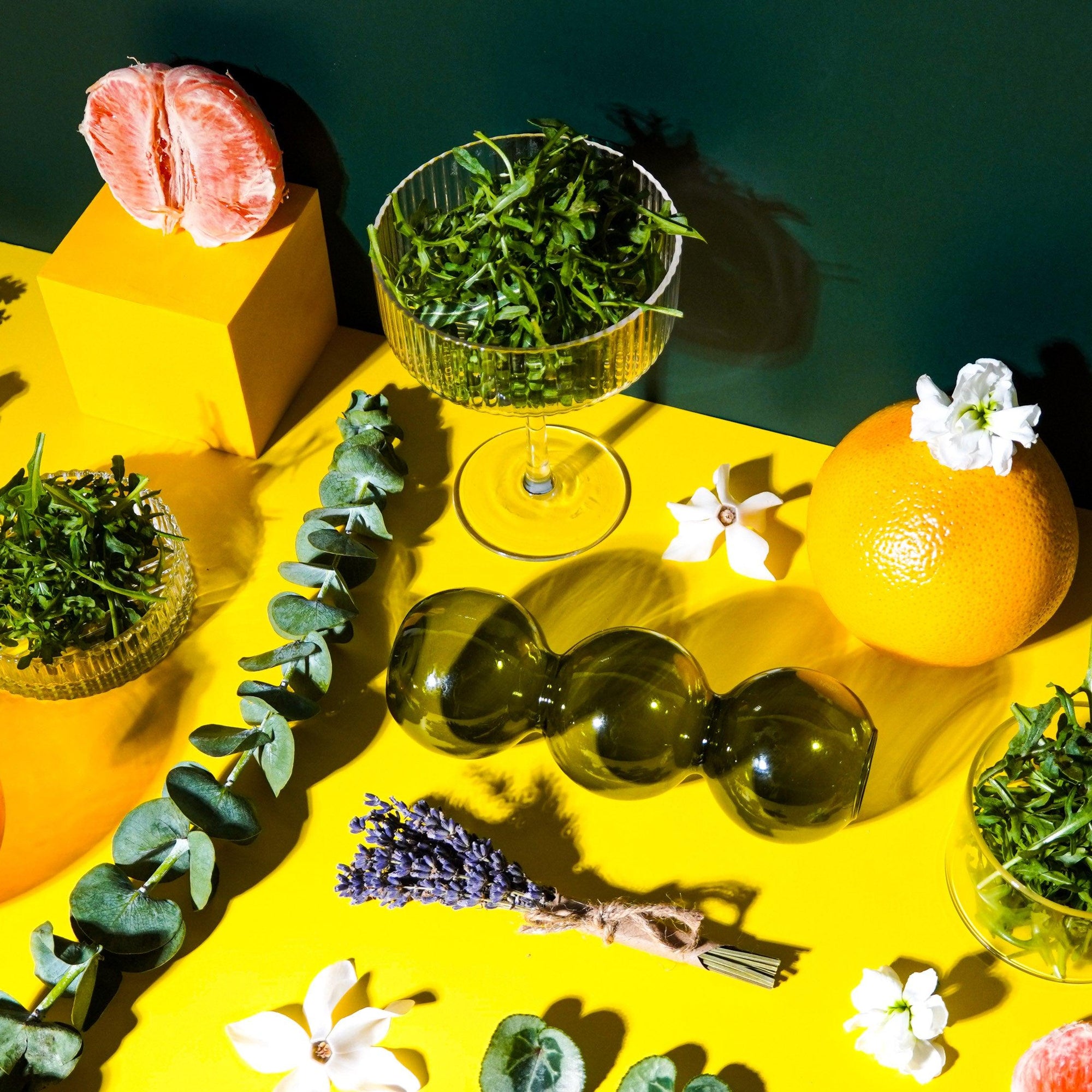A still life arrangement featuring glass bottles, oranges, greenery, and white flowers against a yellow and green background.