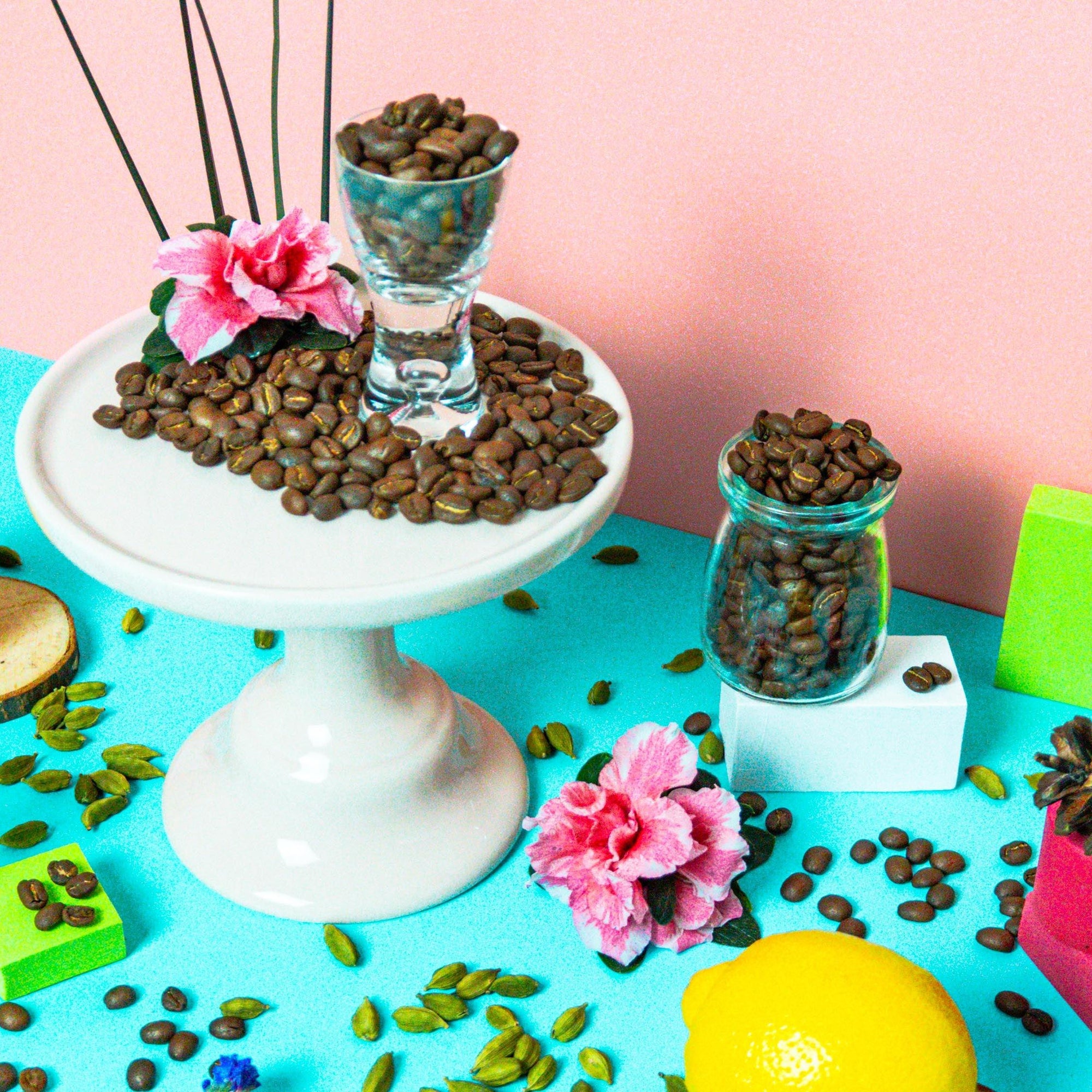 A display featuring a white cake stand with coffee beans on top, surrounded by various props including a clear jar with coffee beans, lemon slices, artificial flowers, and other decorative items against a colorful background.