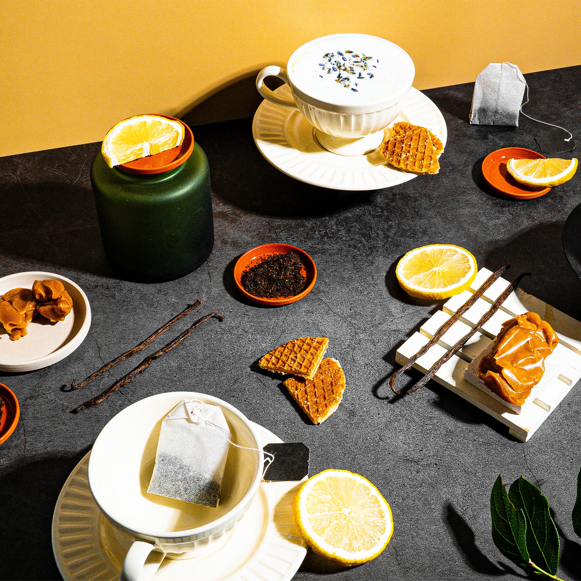 A vibrant, tea-inspired flat lay set against a dark, textured backdrop with a warm golden accent. The scene includes delicate tea cups, one topped with floral garnish and another with marmalade, alongside stroopwafels, lemon slices, caramel swirls, loose tea leaves, and vanilla beans. A green jar adds depth, while sprigs of fresh green leaves bring a natural element to the composition. Perfectly balanced, the setup evokes cozy indulgence with a sophisticated touch of citrus and sweetness.