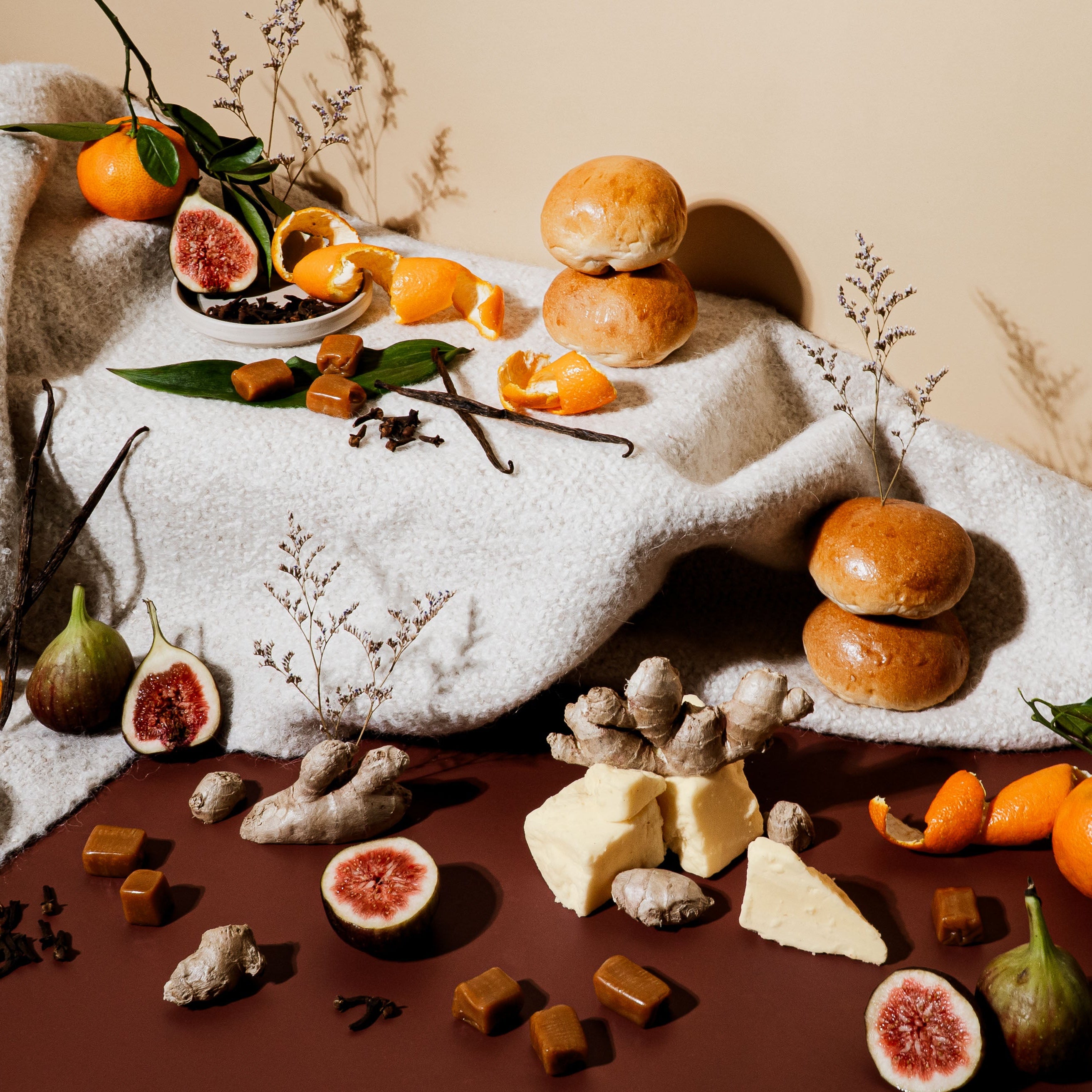 A cozy still life set on a warm brown surface and cream blanket, featuring figs, ginger root, tangerines with peels, clove, vanilla beans, soft rolls, and caramel pieces. The scene is rich with texture and warmth, evoking baked goods, mulled spices, and winter indulgence with a soft glow of comfort.
