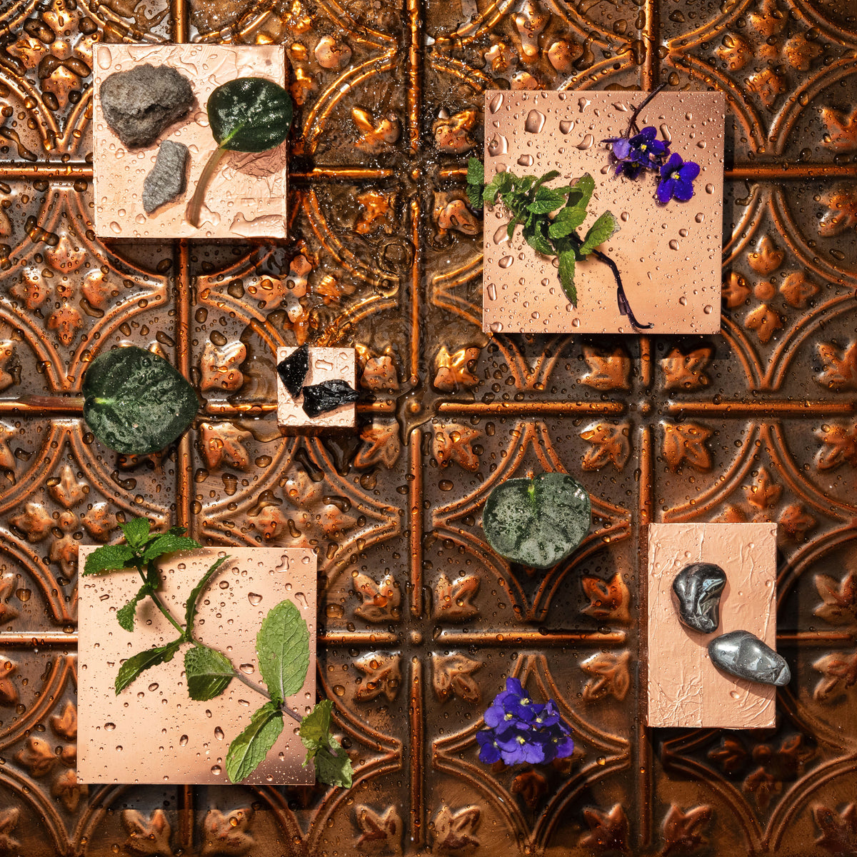Four square copper-coloured tiles arranged on a decorative embossed copper-toned metal surface, each holding small natural stones, green leaves, and purple flowers with water droplets.