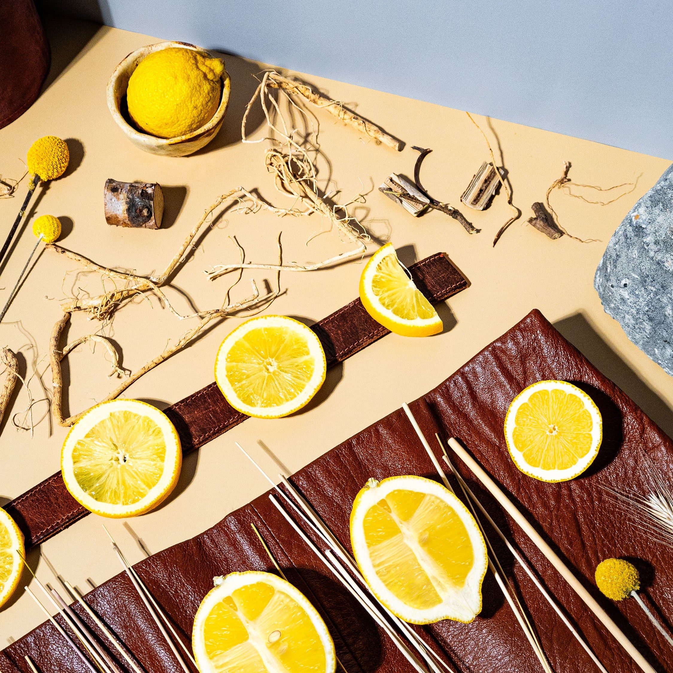 A striking arrangement of bright lemon slices and zesty whole lemons contrasts against rugged leather, earthy roots, and dried botanicals. A modern composition combining natural textures and vibrant citrus, evoking freshness with a touch of rustic sophistication.