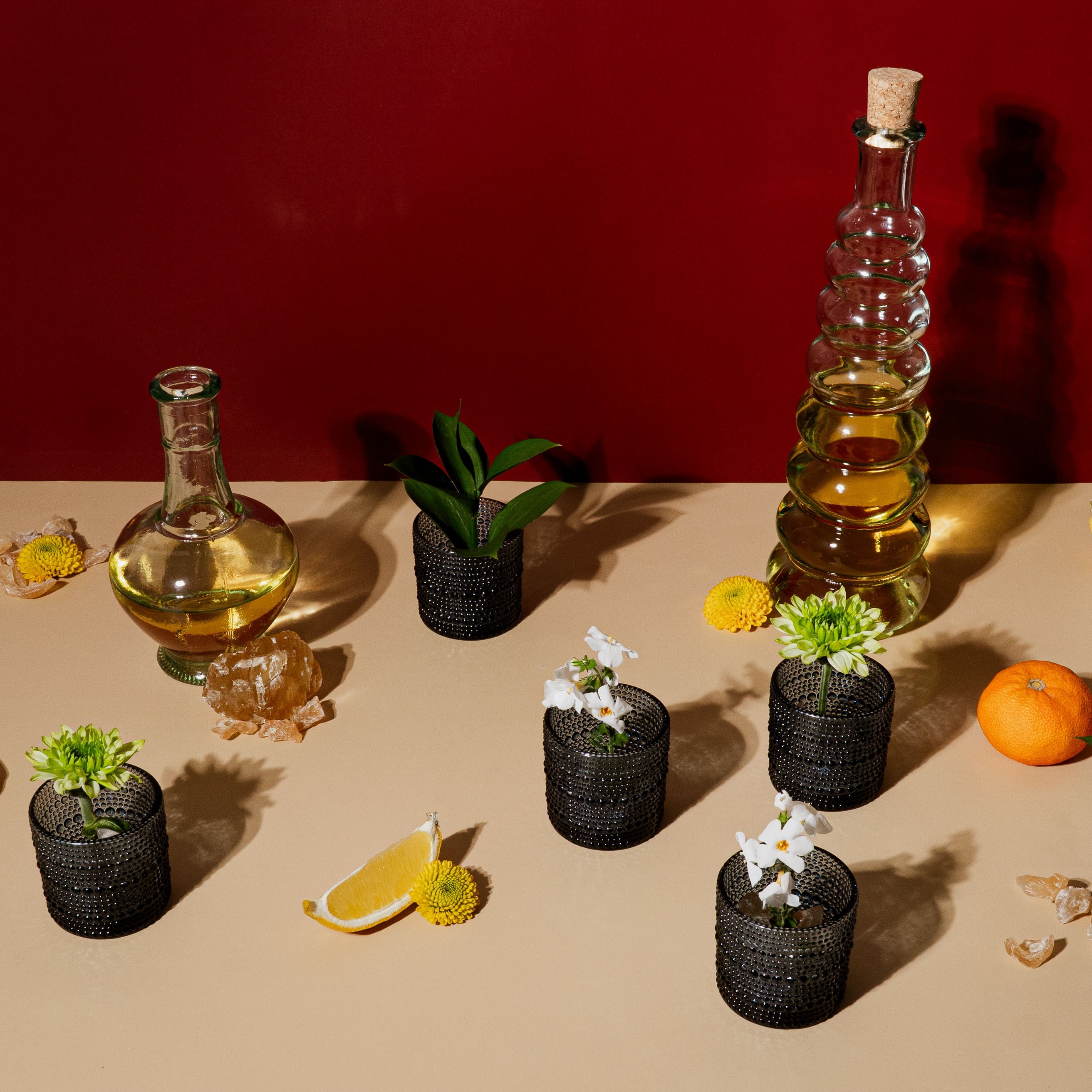 A still life on a beige surface with a deep red backdrop, featuring glass bottles filled with golden oil, citrus fruits, lemon wedges, fresh greenery, and small black textured vases holding white and yellow flowers. Amber resin pieces and slices of lemon and orange are scattered around, evoking a warm, radiant mood inspired by ritual and light.