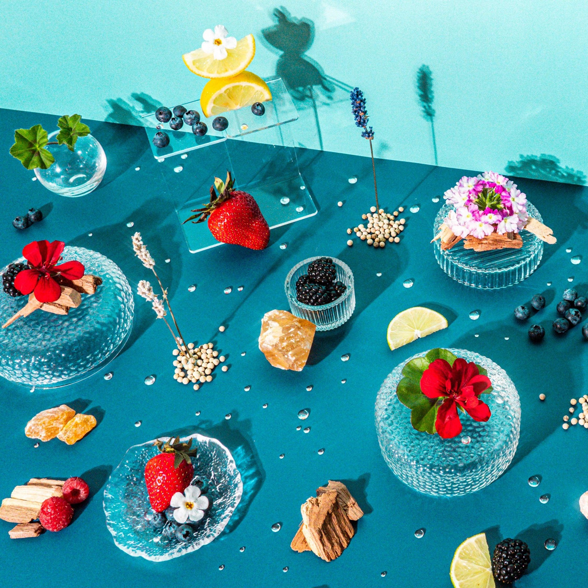 A delightful and artistic display of botanical and culinary elements on a teal background. The scene includes various fresh berries like blueberries and strawberries, slices of lemon, and vibrant flowers, all artistically arranged on textured glass plates and alongside pieces of wood and dried herbs. The composition is accentuated by glass vases holding sprigs of lavender and other dried botanicals, creating a fresh, vibrant tableau that evokes a sense of summer freshness and natural beauty.