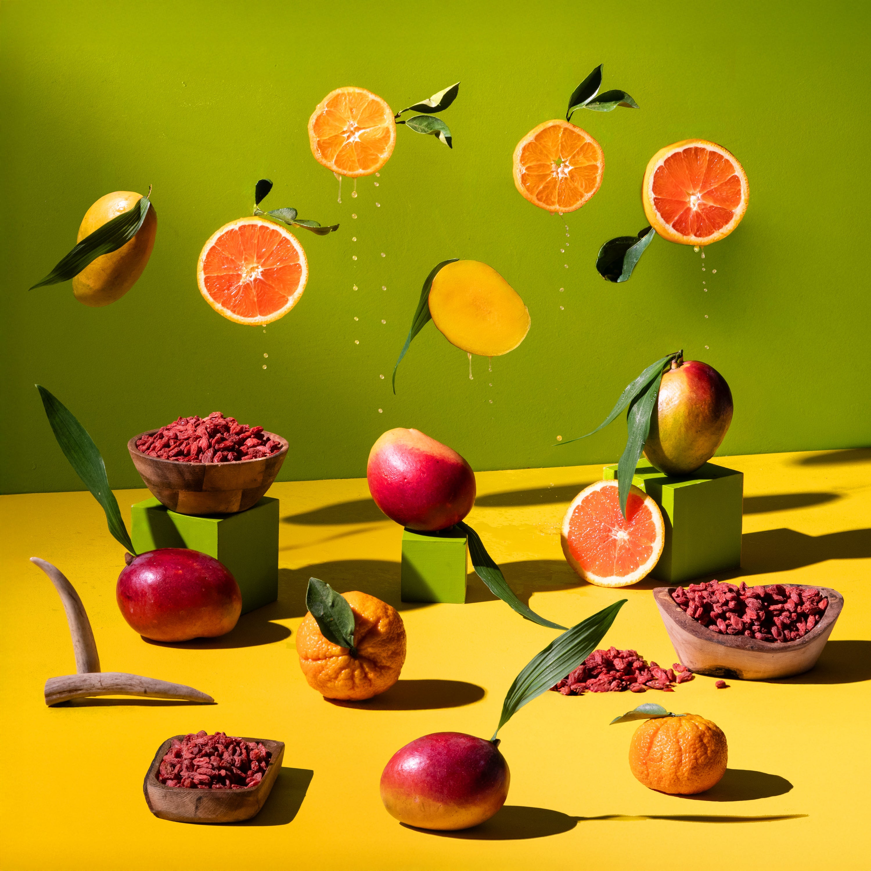 Bright still-life arrangement of goji berries, mangoes, citrus slices, leaves, and wooden bowls on a yellow surface against a green background, with sharp sunlight and bold shadows.