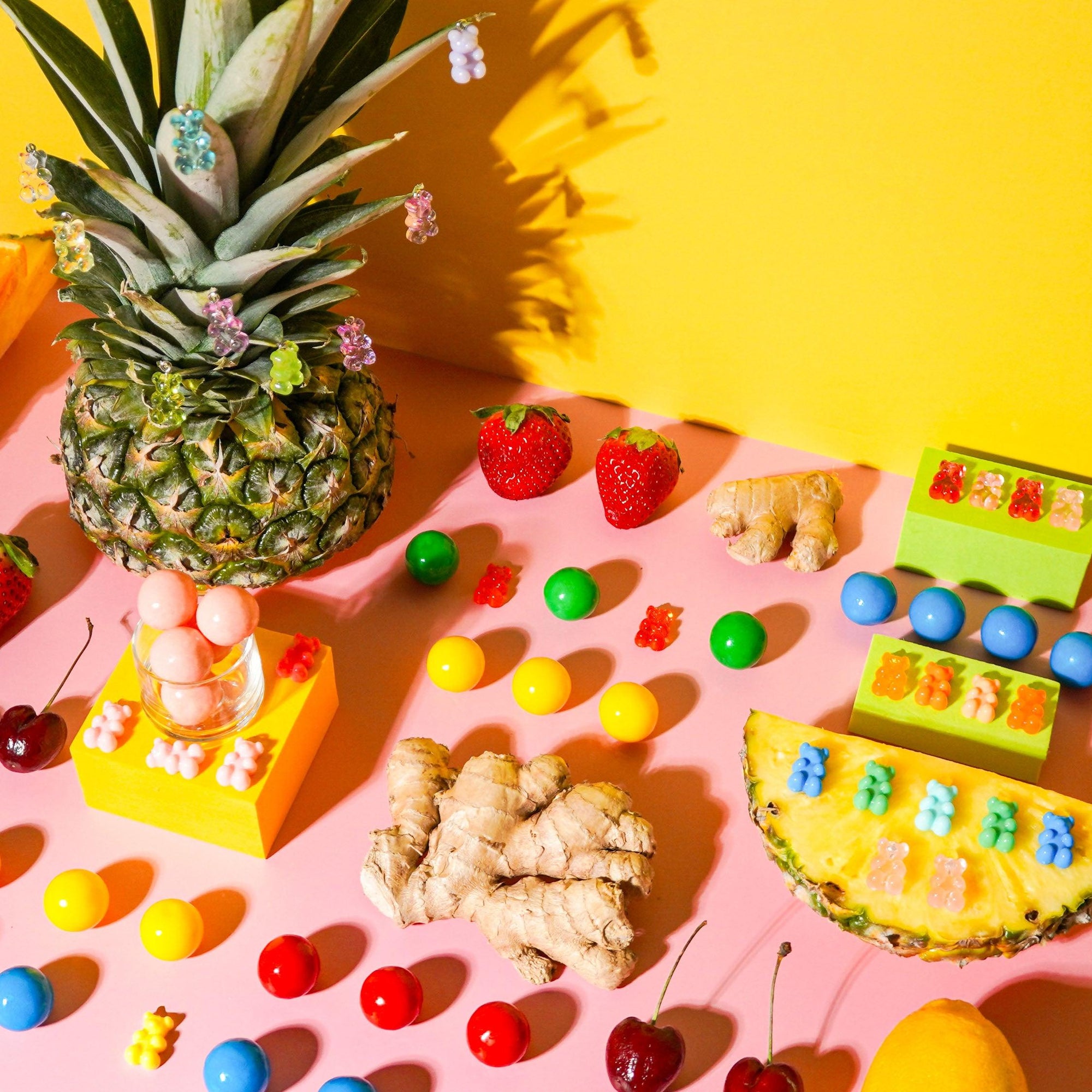 A colorful assortment of fragrance oils presented in small containers, arranged alongside fruit and candy-like items on a yellow and pink background. A pineapple, strawberries, an orange, and various sweets are visible.