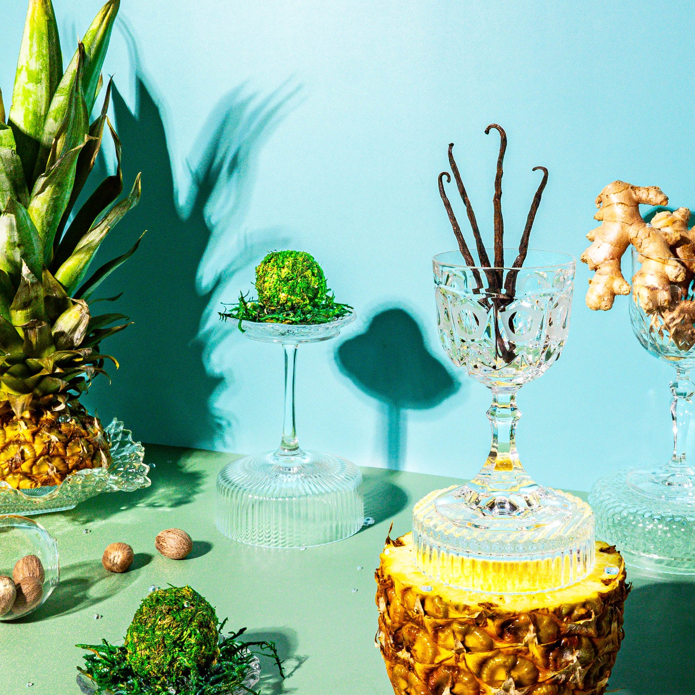 A vibrant tropical display with a fresh pineapple, vanilla beans, ginger roots, moss spheres, and scattered nutmeg seeds, arranged in elegant crystal glassware against a turquoise background.