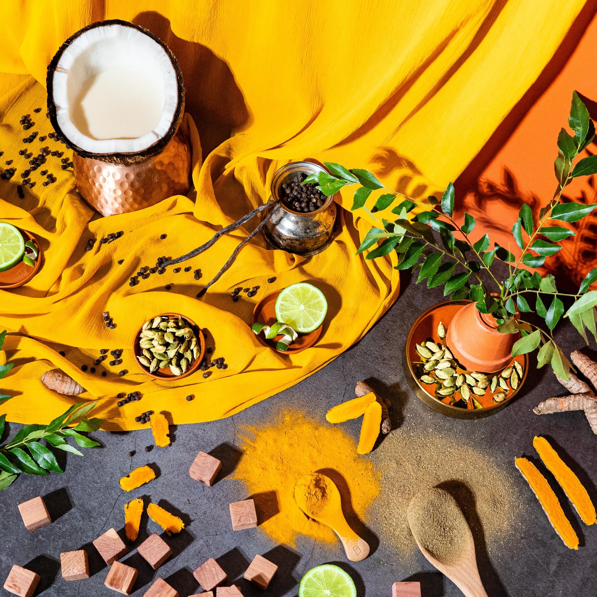 Vibrant flat lay, for fragrance oil, inspired by South Asian ingredients featuring coconut, cardamom pods, black peppercorns, turmeric roots and powder, curry leaves, lime halves, wooden cubes, and a draped yellow fabric on a dark stone surface with an orange backdrop