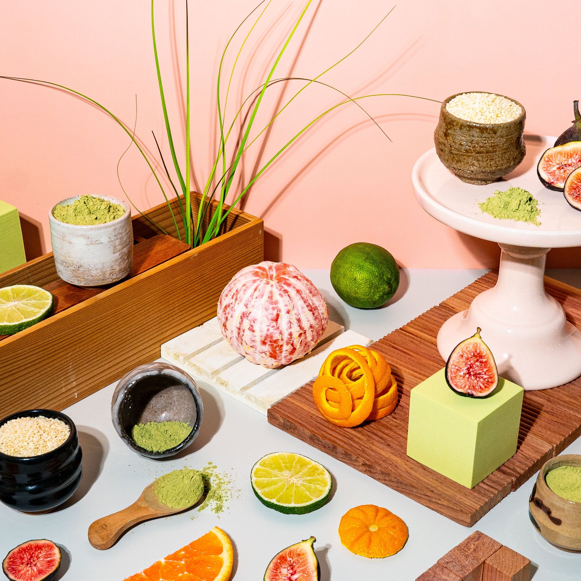 A vibrant, carefully arranged still life featuring fresh figs, citrus slices, matcha powder, and grains against a warm peach toned backdrop. Earthy matcha and bright limes sit alongside rich figs and fragrant orange peels, creating a scene that feels both natural and curated. Ceramic bowls and wooden surfaces add texture, evoking the balance of freshness and warmth. The composition is playful yet refined, celebrating the depth of scent and flavour in each element.