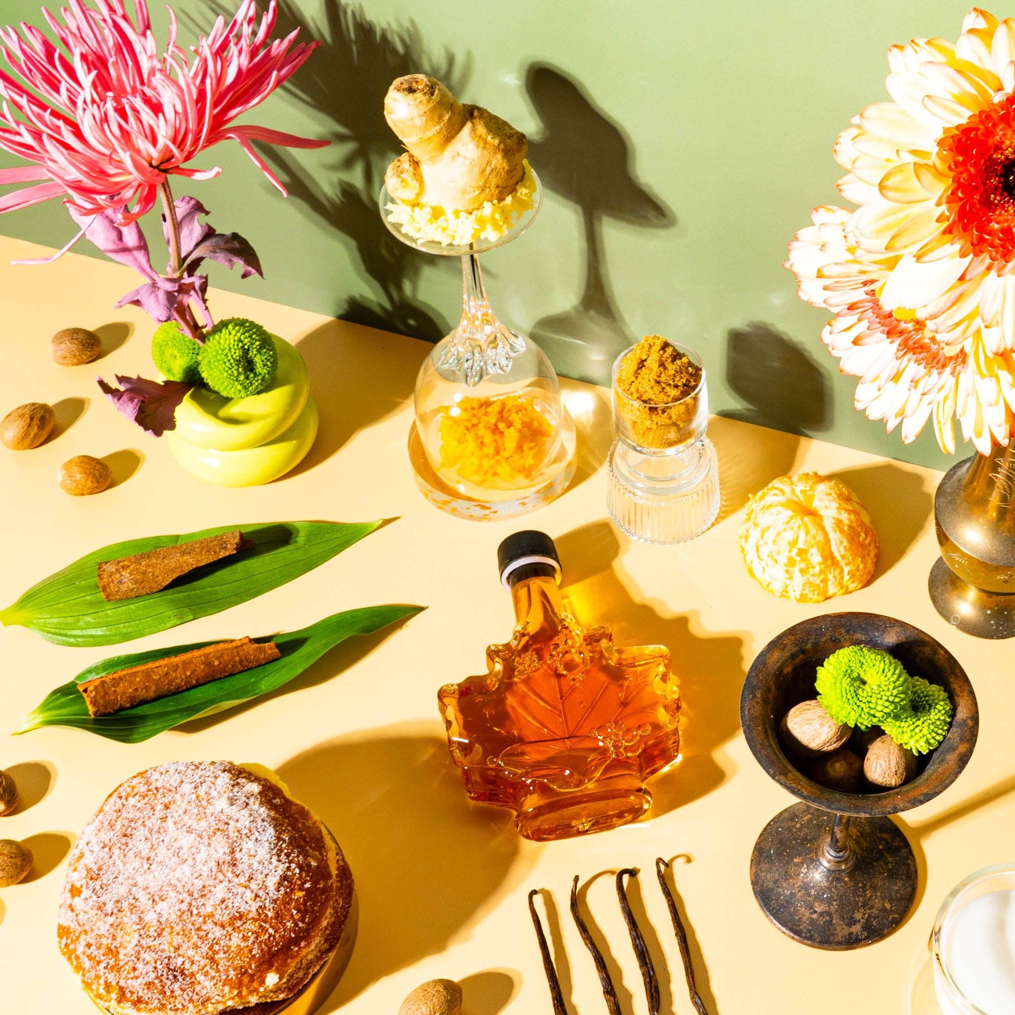 An assortment of items including a bottle of maple syrup, a bowl of brown sugar, grated ginger, a buttermilk container, and various baking ingredients arranged on a table with a green background. Floral decorations are also visible in the corners of the image.