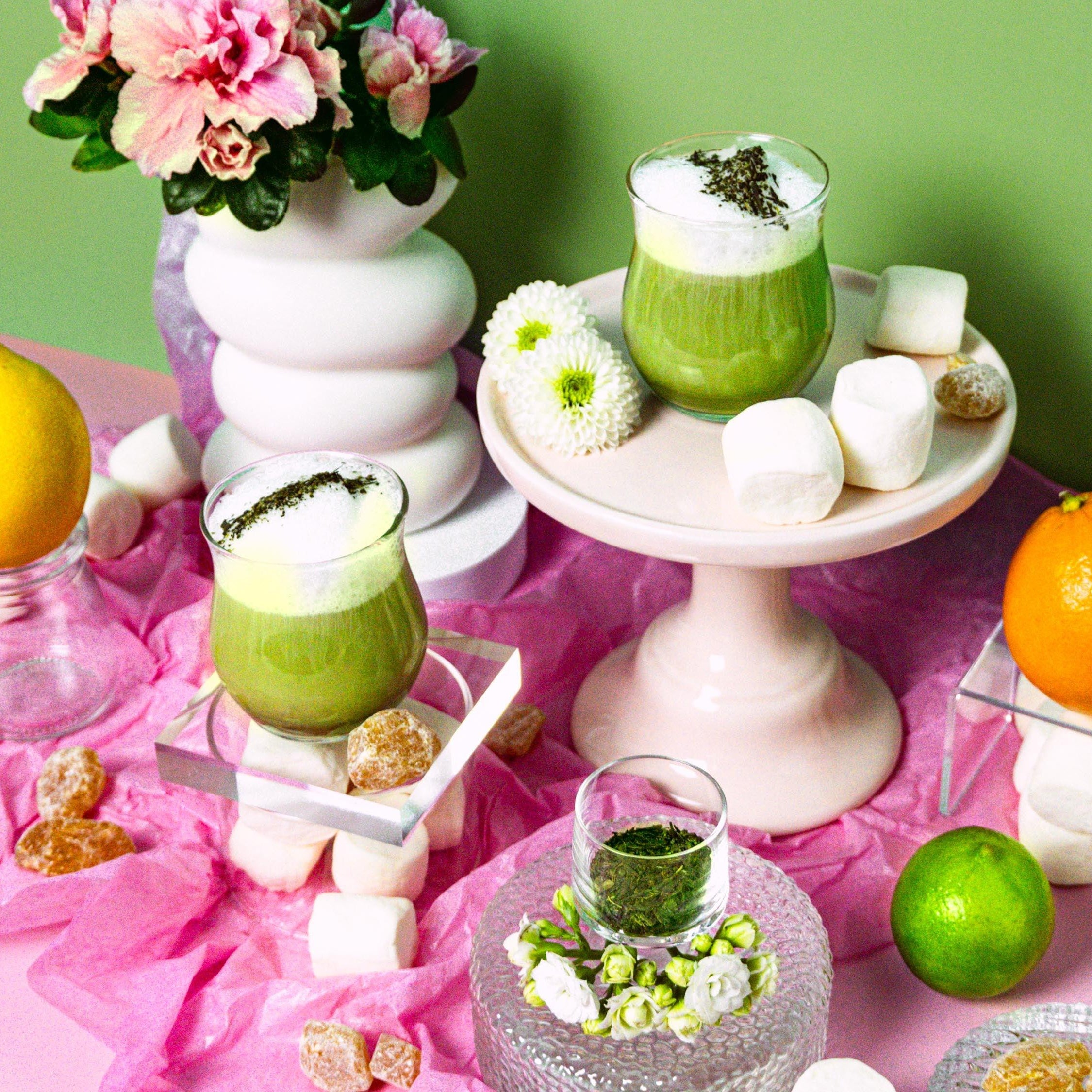 A display featuring fragrance oil in a green container with decorative elements including lemon, orange, and marshmallows, presented on a pink surface.
