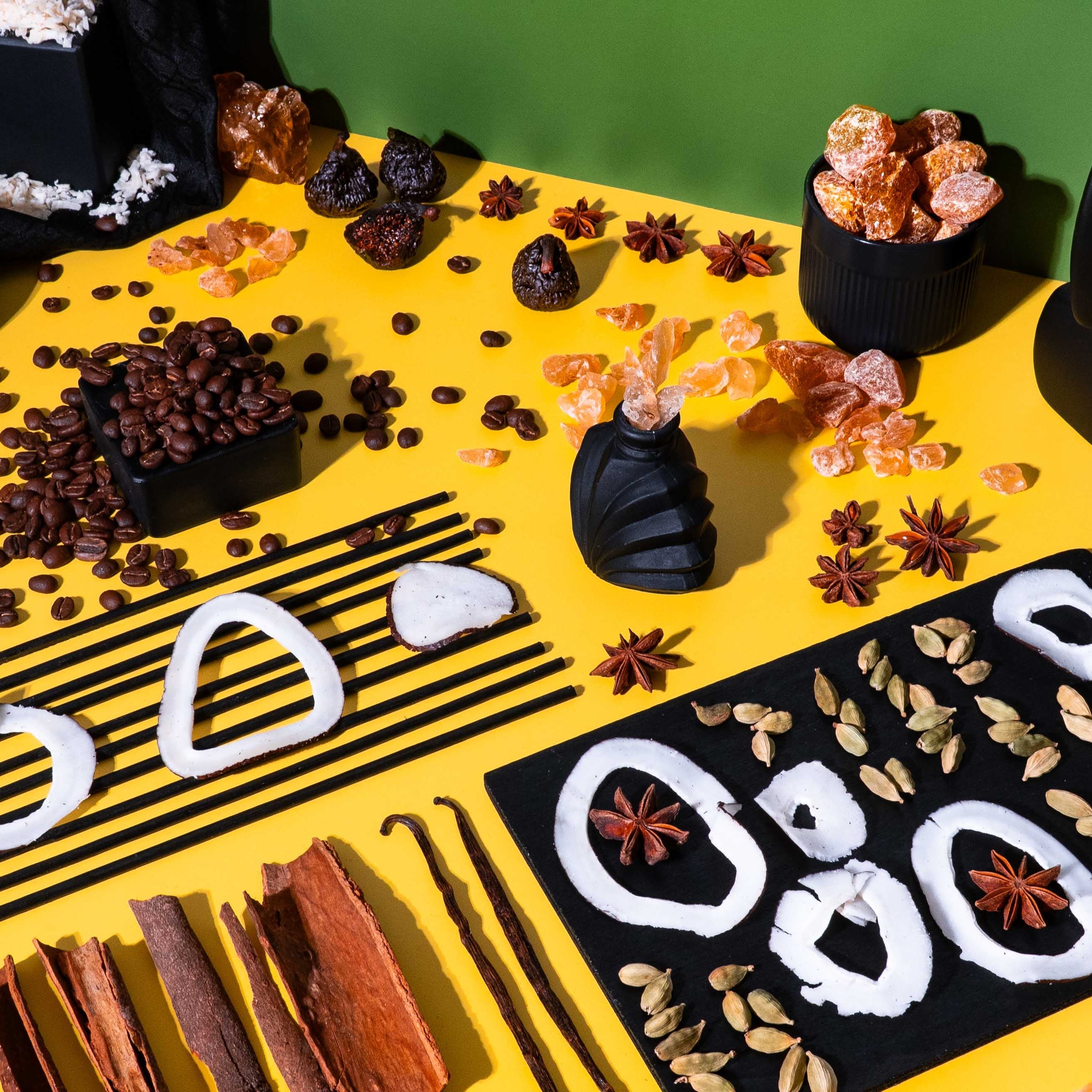 Flat lay of fragrance oil ingredients on a split yellow and green background featuring coffee beans, coconut rings, cardamom pods, cinnamon sticks, star anise, vanilla beans, dried figs, and rock sugar, arranged in a modern, graphic composition with black ceramic vessels and abstract shapes.