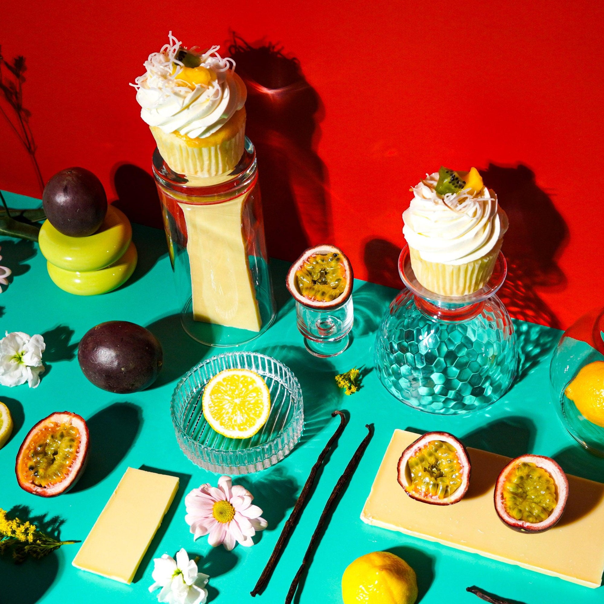 A table setting with passion fruit cupcakes topped with white chocolate ganache, presented with passion fruit halves, lemons, and decorative flowers against a red and turquoise background.