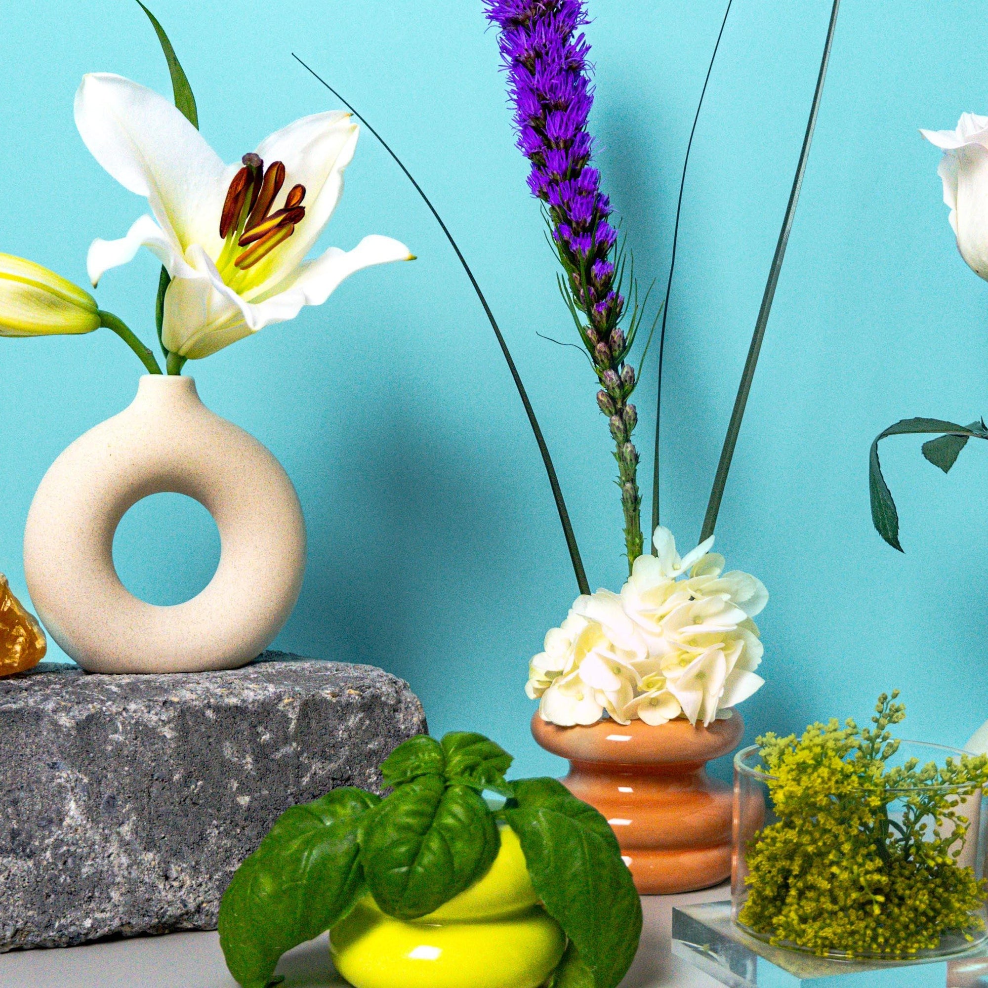 A composition featuring a white T-shirt with a fragrance oil bottle, surrounded by various flowers in different vases, against a blue background.