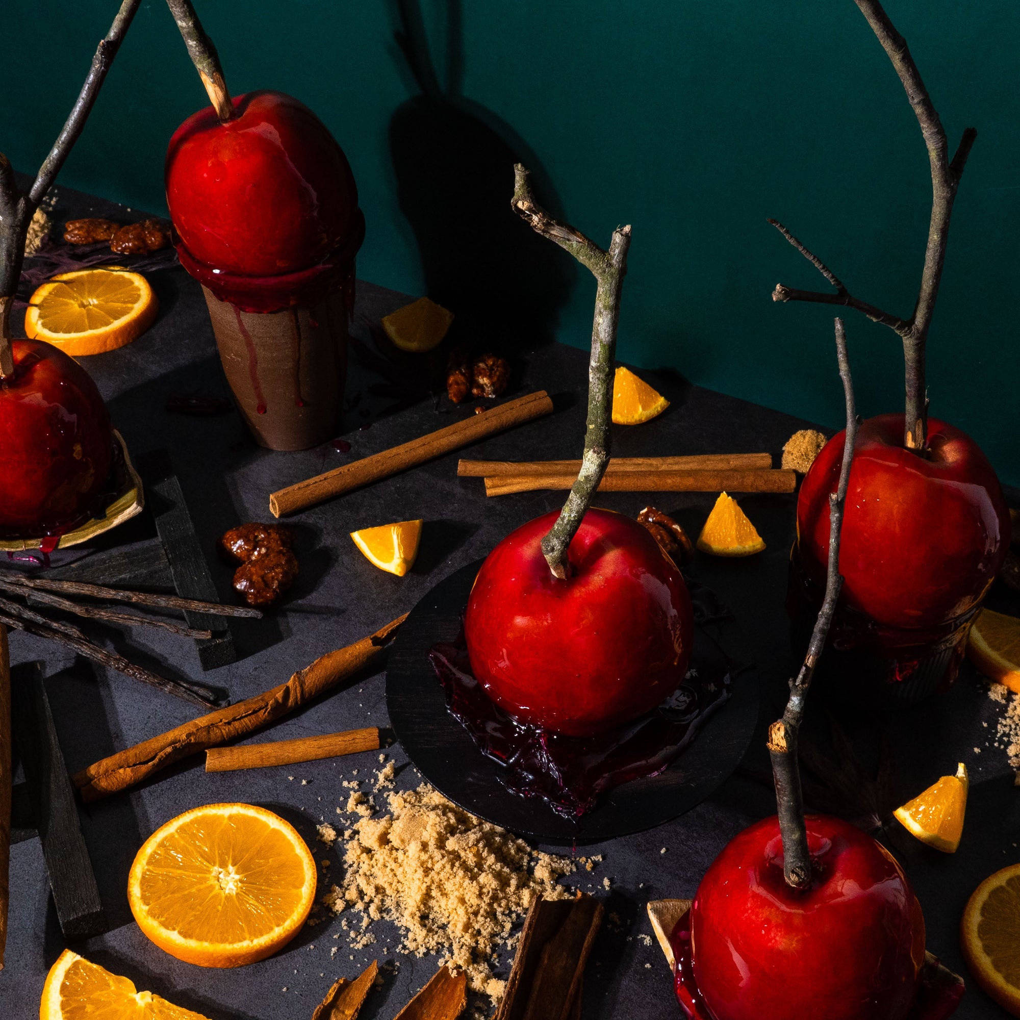 Cinematic flat lay for a fragrance oil, of candy apples on dark surface with cinnamon sticks, brown sugar, orange slices, and vanilla beans, styled with rustic twigs and deep shadows. Autumn carnival-inspired fragrance photography.