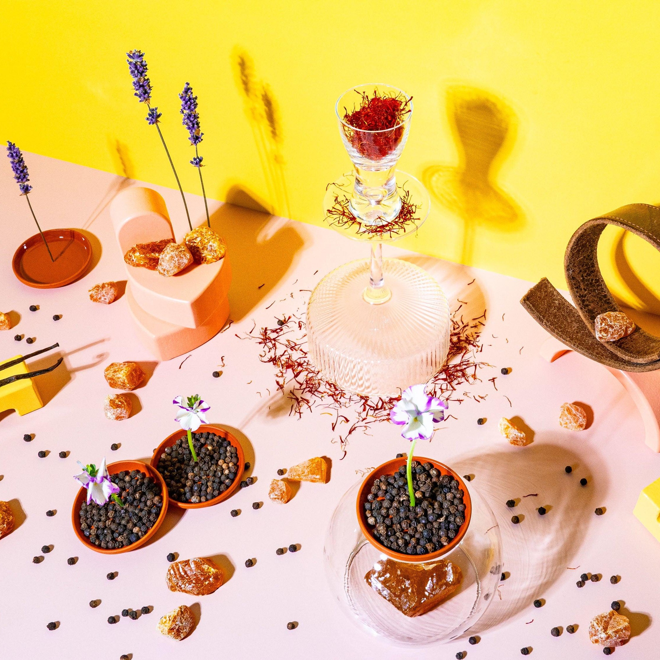 A vibrant and artistic tabletop display on a yellow background featuring a variety of objects: small terracotta pots with black peppercorns, delicate purple flowers, geometrically shaped wooden sculptures, and amber-colored rocks scattered across a pink surface. In the center, a clear glass filled with vibrant red spices casts playful shadows, accompanied by lavender stalks adding a touch of floral elegance. The scene is carefully arranged to create a visually stimulating and colourful composition.
