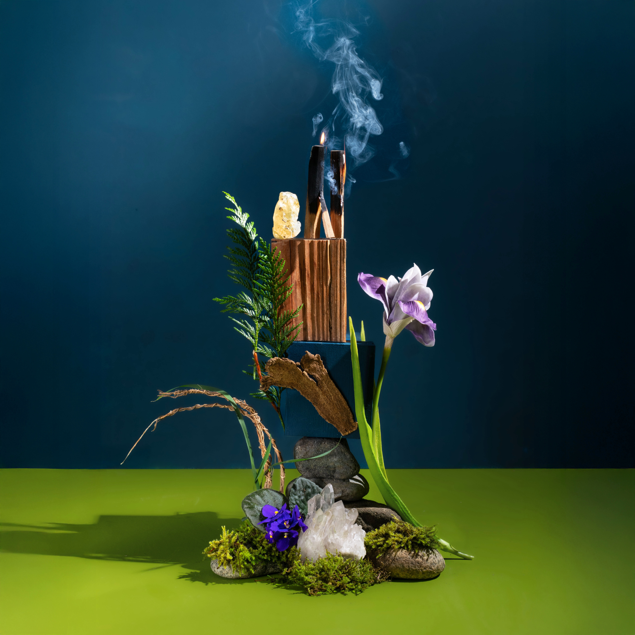 Decorative setup with incense sticks, flowers, and stones on a green surface against a dark background
