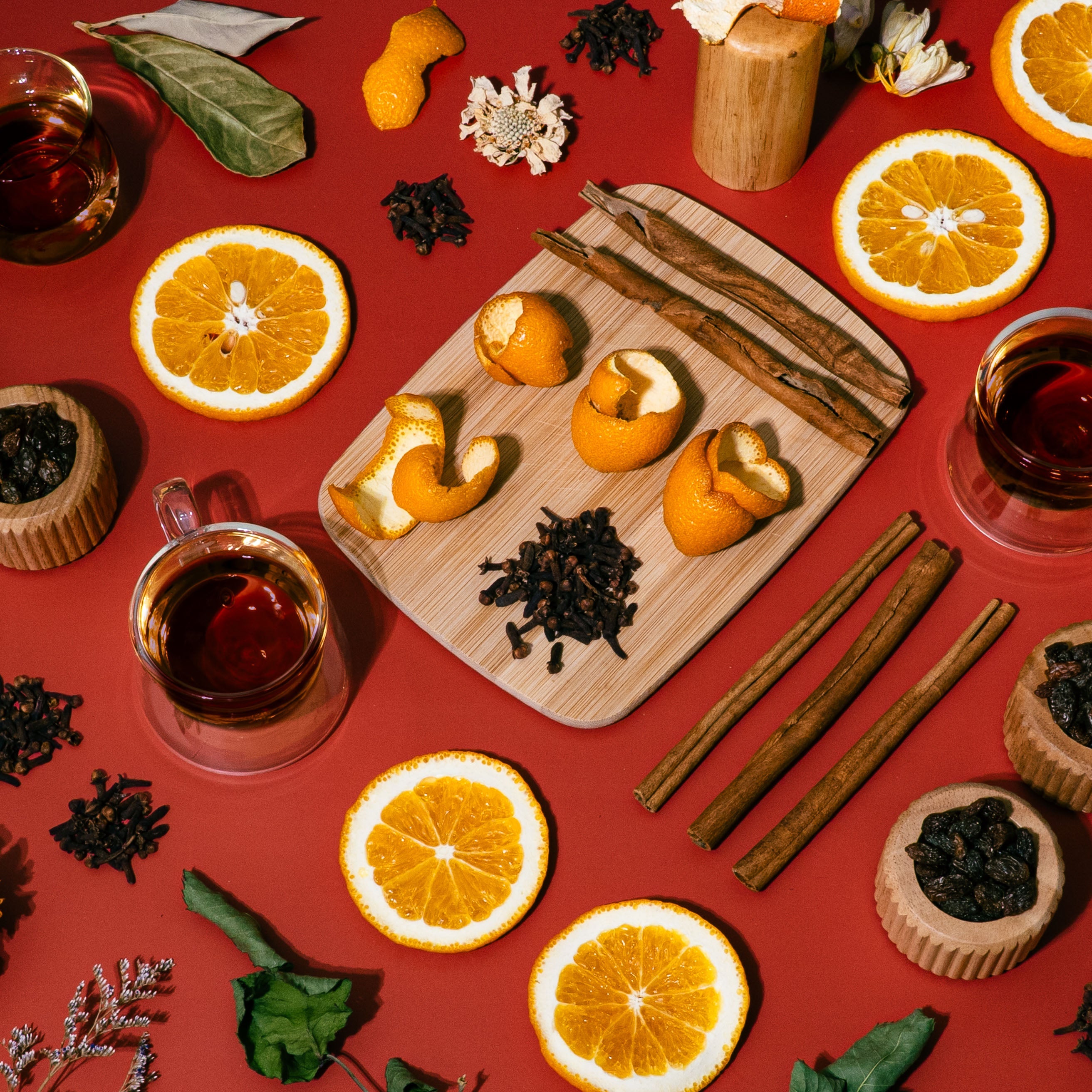 A warm toned flat lay on a rich red background featuring dried orange slices, cinnamon sticks, clove, orange peels, raisins in paper cups, bay leaves, mugs of spiced tea, and dried florals. The scene evokes a cozy, spiced autumn or winter gathering with strong sensory cues of citrus, warmth, and seasonal tradition.