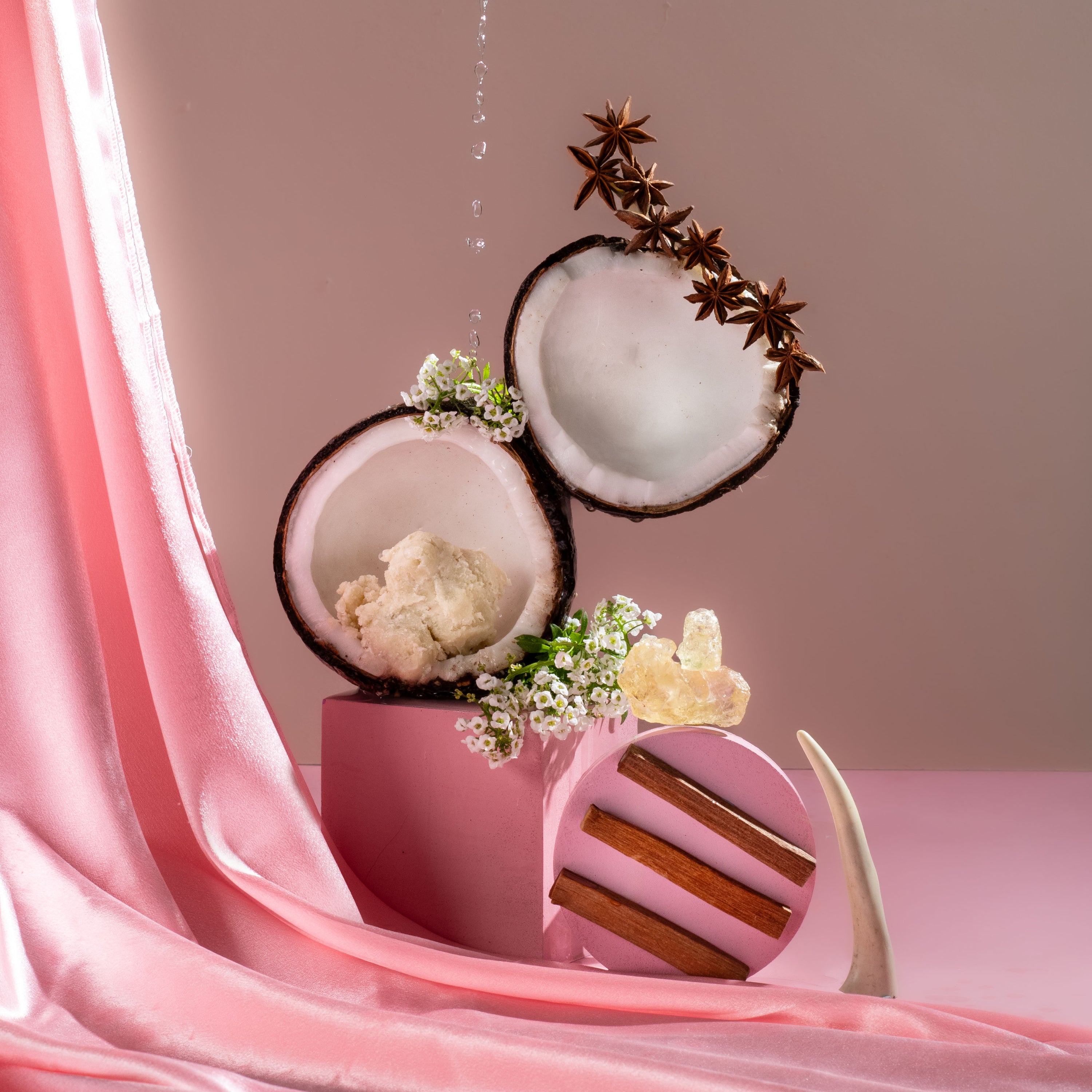 Decorative setup with coconut halves, flowers, and a pink container on a pink fabric background.