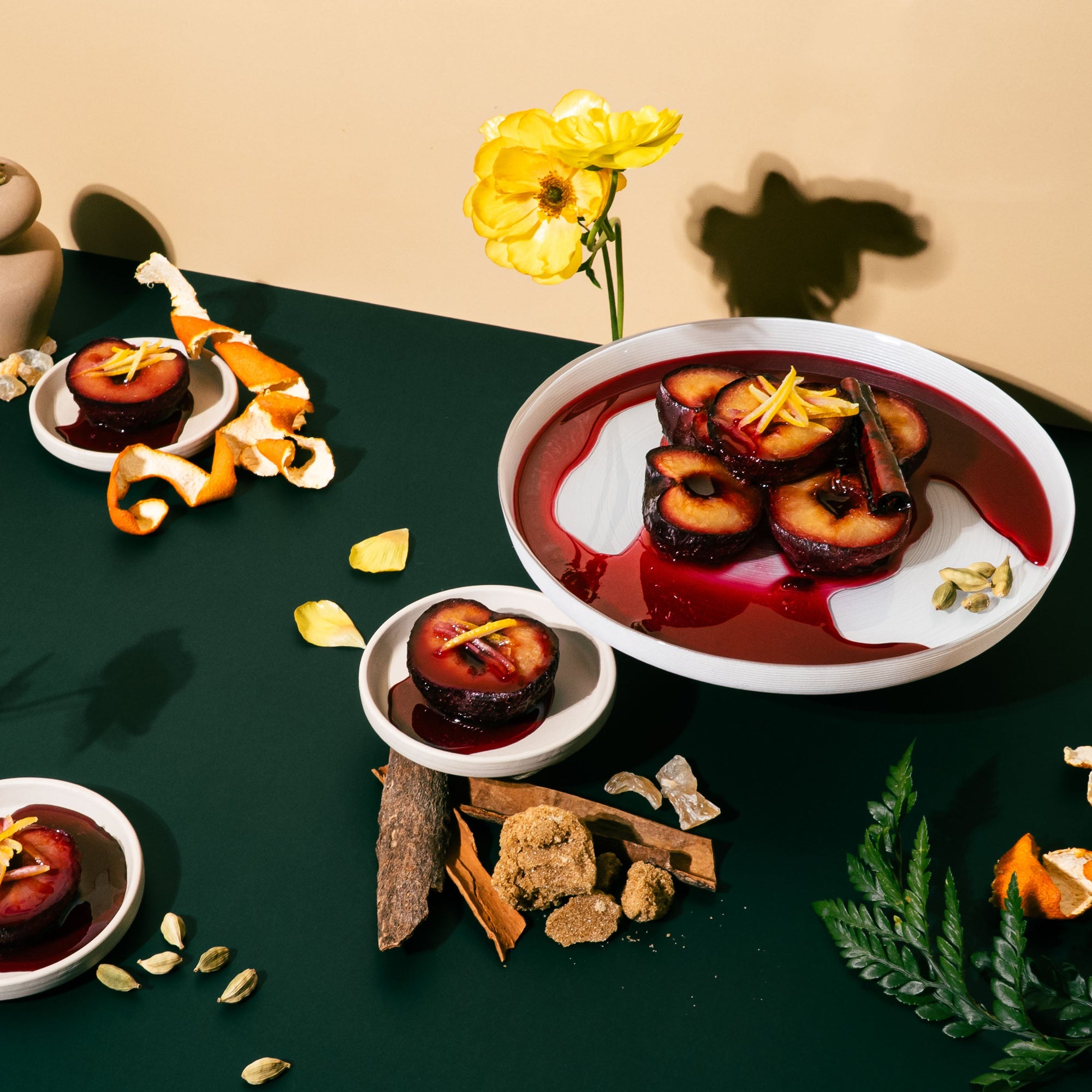 A dramatic still life on a dark green surface featuring halved plums glazed in red syrup, garnished with citrus peel and cardamom. The scene includes bowls of poached plums, cinnamon bark, orange peels, yellow flowers, brown sugar, walnuts, and greenery, evoking a rich, spiced dessert with a deep, sensual warmth.