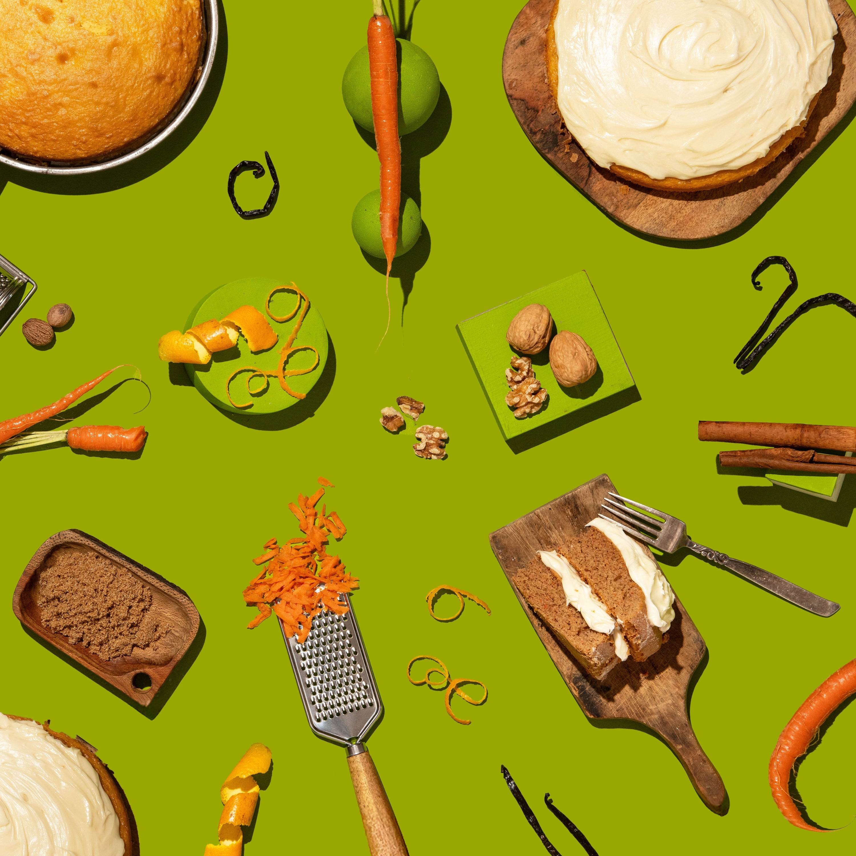 A styled flat-lay of carrot cake ingredients and slices arranged on a bright green background, including cake pieces with white frosting, carrots, walnuts, cinnamon sticks, a grater, and orange zest.