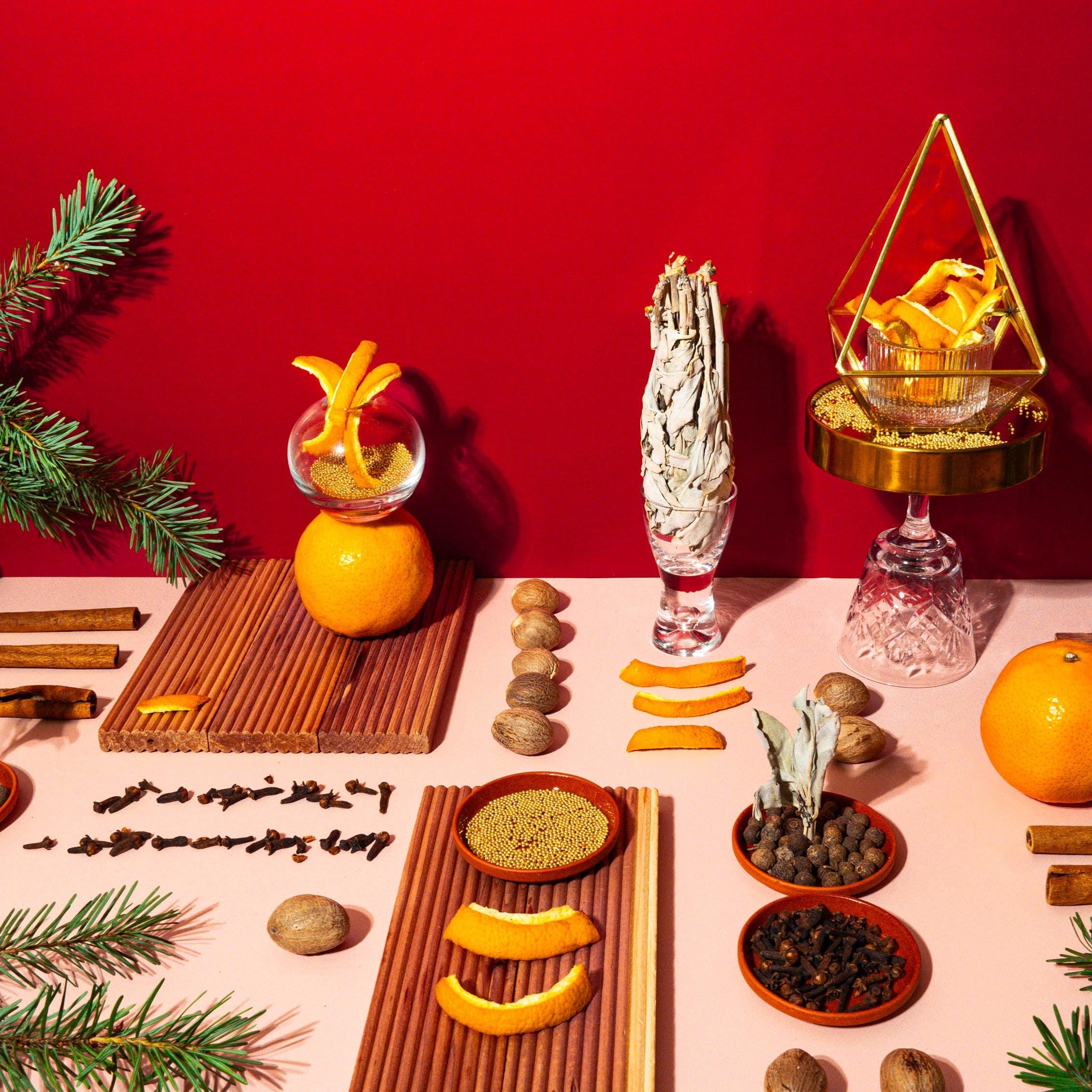 A vibrant holiday tablescape featuring an array of festive elements on a red background. Central to the image is a display of fresh and dried oranges, cinnamon sticks, cloves, and nutmeg, alongside evergreen branches and a pyramid-shaped terrarium filled with golden accents. On the left, an orange is adorned with a peel spiral standing on a wooden board, adding a touch of whimsy. Two elegant glassware pieces complete the scene, evoking a warm, celebratory mood reminiscent of Christmas spices and traditions