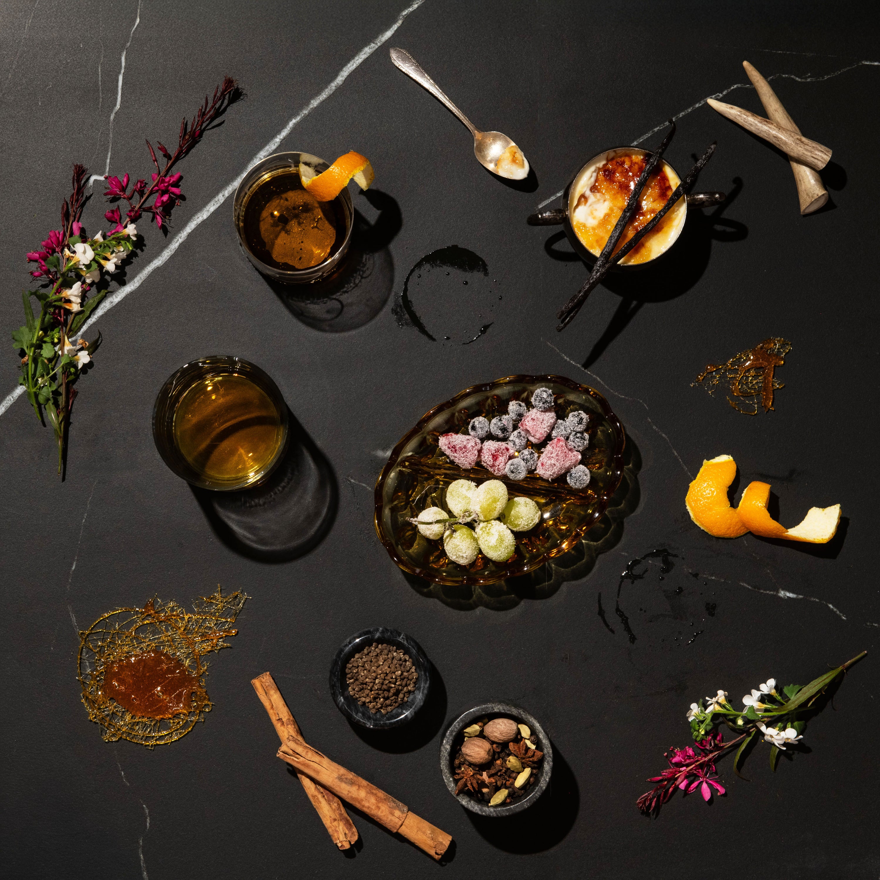 An overhead styled flat-lay of a dark stone surface featuring a central glass bowl of assorted berries and sweets, two glasses of amber liquid, spices, flowers, citrus peels, and small bowls arranged around it.