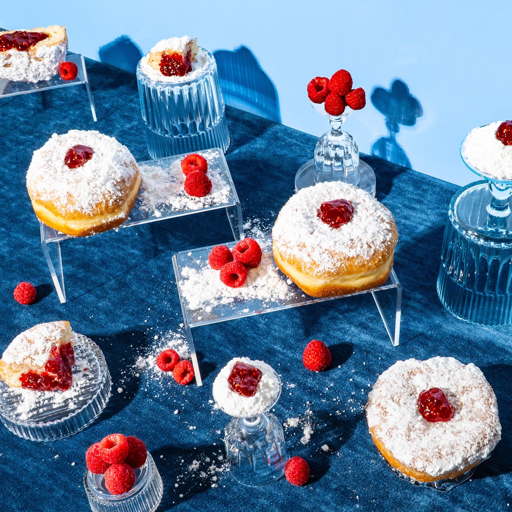 Bright flat lay for a fragrance oil, featuring powdered jelly-filled donuts, fresh raspberries, and glassware on a rich blue fabric background with a soft blue wall, styled with powdered sugar and acrylic risers for a playful, modern dessert scene.