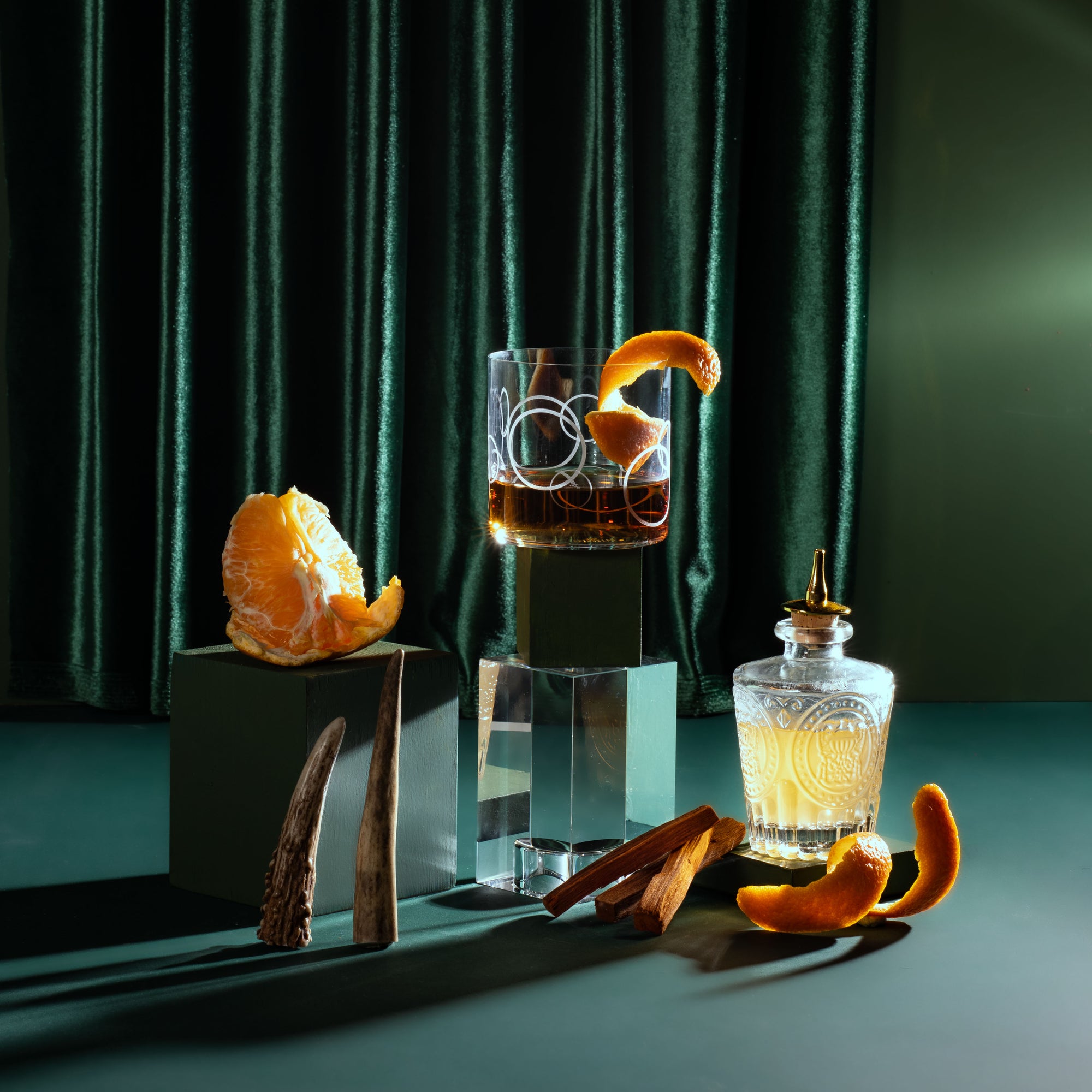 Cocktail with orange garnish and a bottle of liquor on a dark green background
