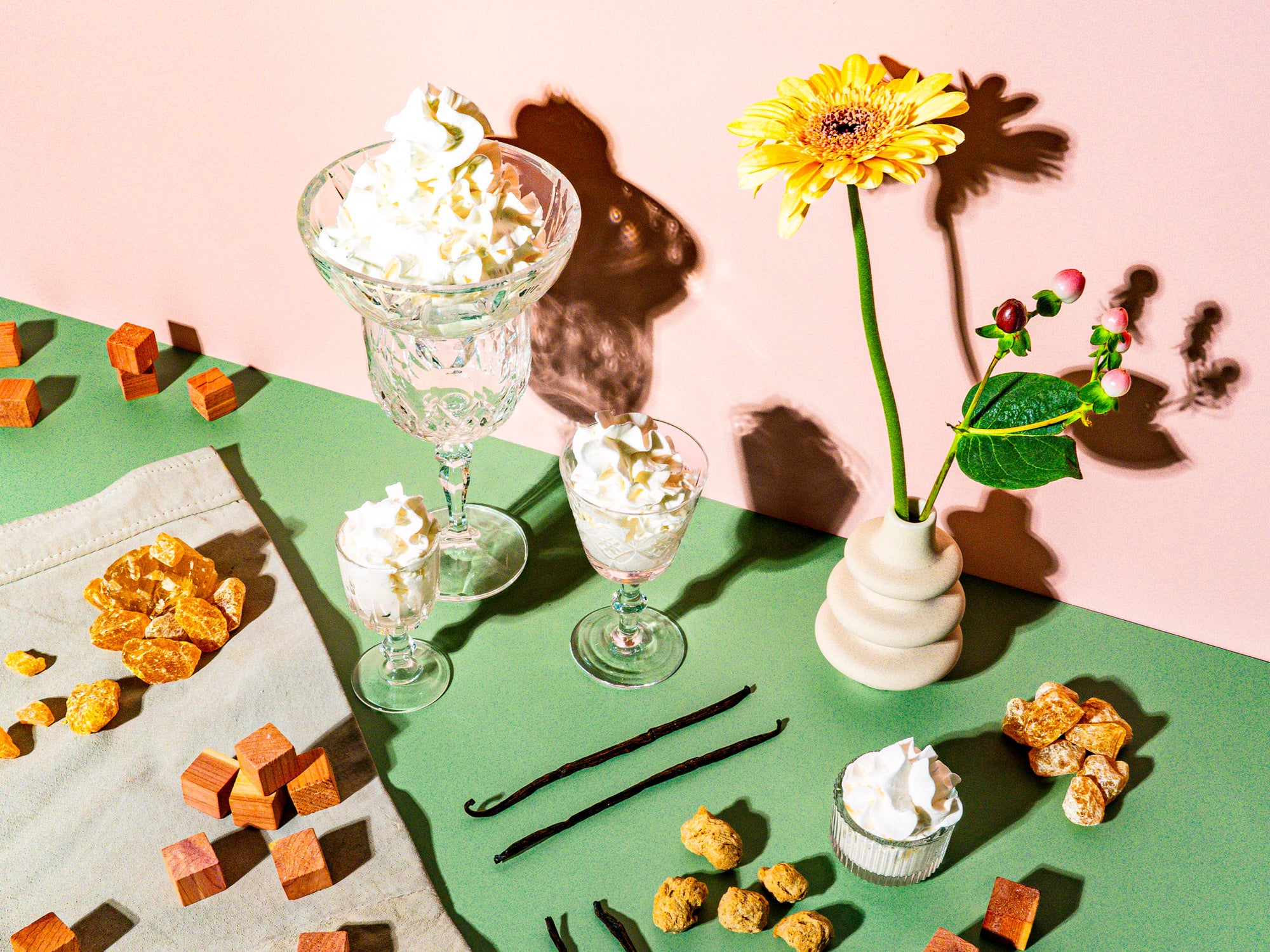 A whimsical, dessert-inspired arrangement featuring whipped cream delicately swirled in crystal glasses, set against a soft pink and green backdrop. The table is adorned with vanilla beans, amber-like caramel shards, wood cubes, and textured nuggets, evoking warmth and sweetness. A vibrant yellow gerbera daisy in a modern vase, paired with red berry accents, adds a natural yet playful touch. The scene radiates creamy decadence with a sophisticated, earthy balance, creating a visual feast of textures