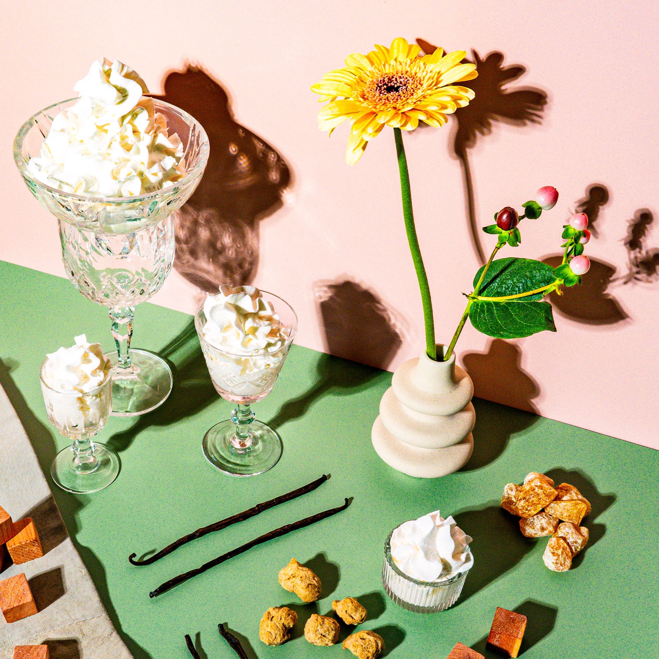 A whimsical, dessert-inspired arrangement featuring whipped cream delicately swirled in crystal glasses, set against a soft pink and green backdrop. The table is adorned with vanilla beans, amber-like caramel shards, wood cubes, and textured nuggets, evoking warmth and sweetness. A vibrant yellow gerbera daisy in a modern vase, paired with red berry accents, adds a natural yet playful touch. The scene radiates creamy decadence with a sophisticated, earthy balance, creating a visual feast of textures