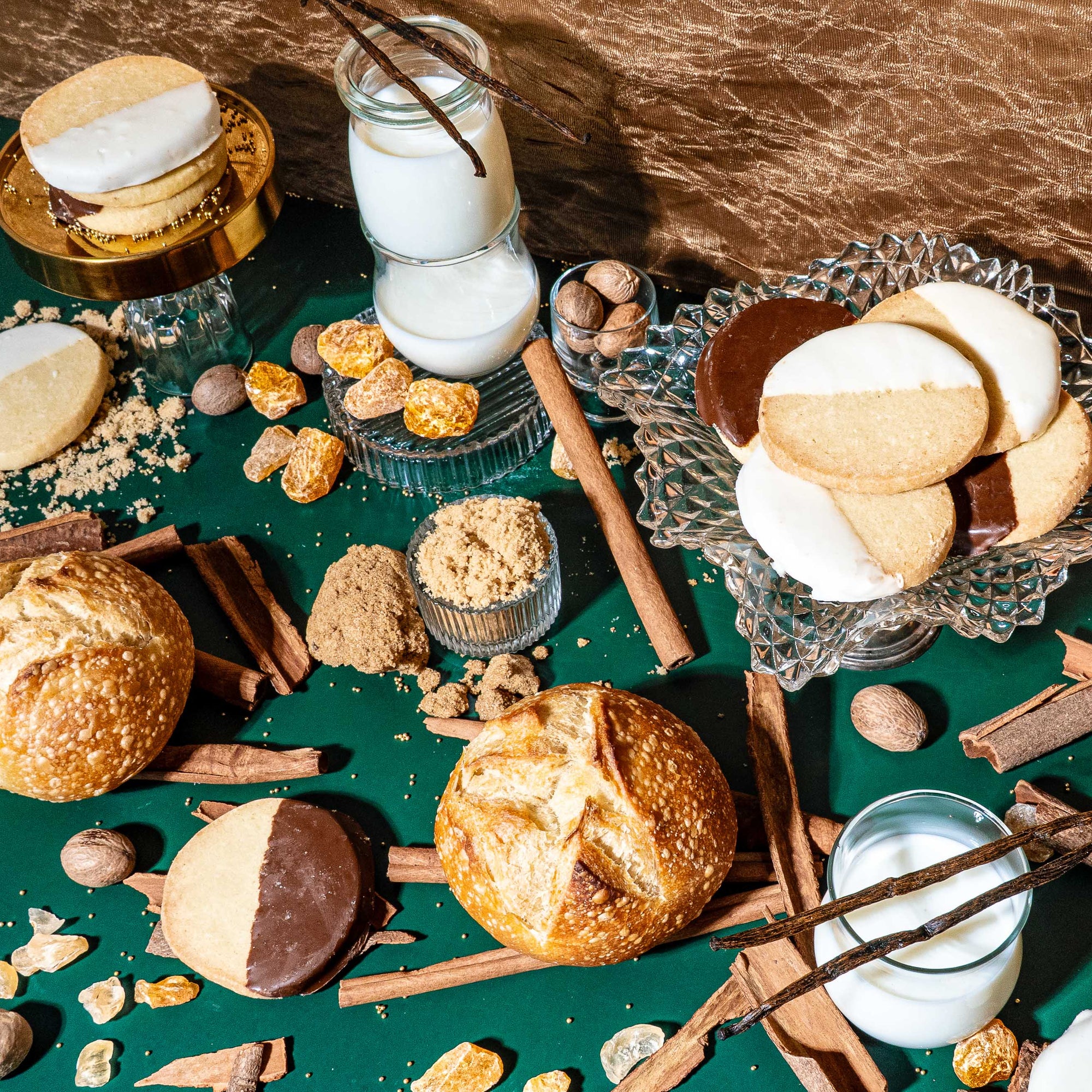 A festive, indulgent flat lay featuring black-and-white cookies, crusty bread rolls, glasses of milk, cinnamon sticks, vanilla beans, nutmeg, brown sugar, and shards of golden brittle. Set against a deep green tablecloth and gold textured backdrop, the scene evokes cozy holiday baking with a rich, nostalgic warmth.