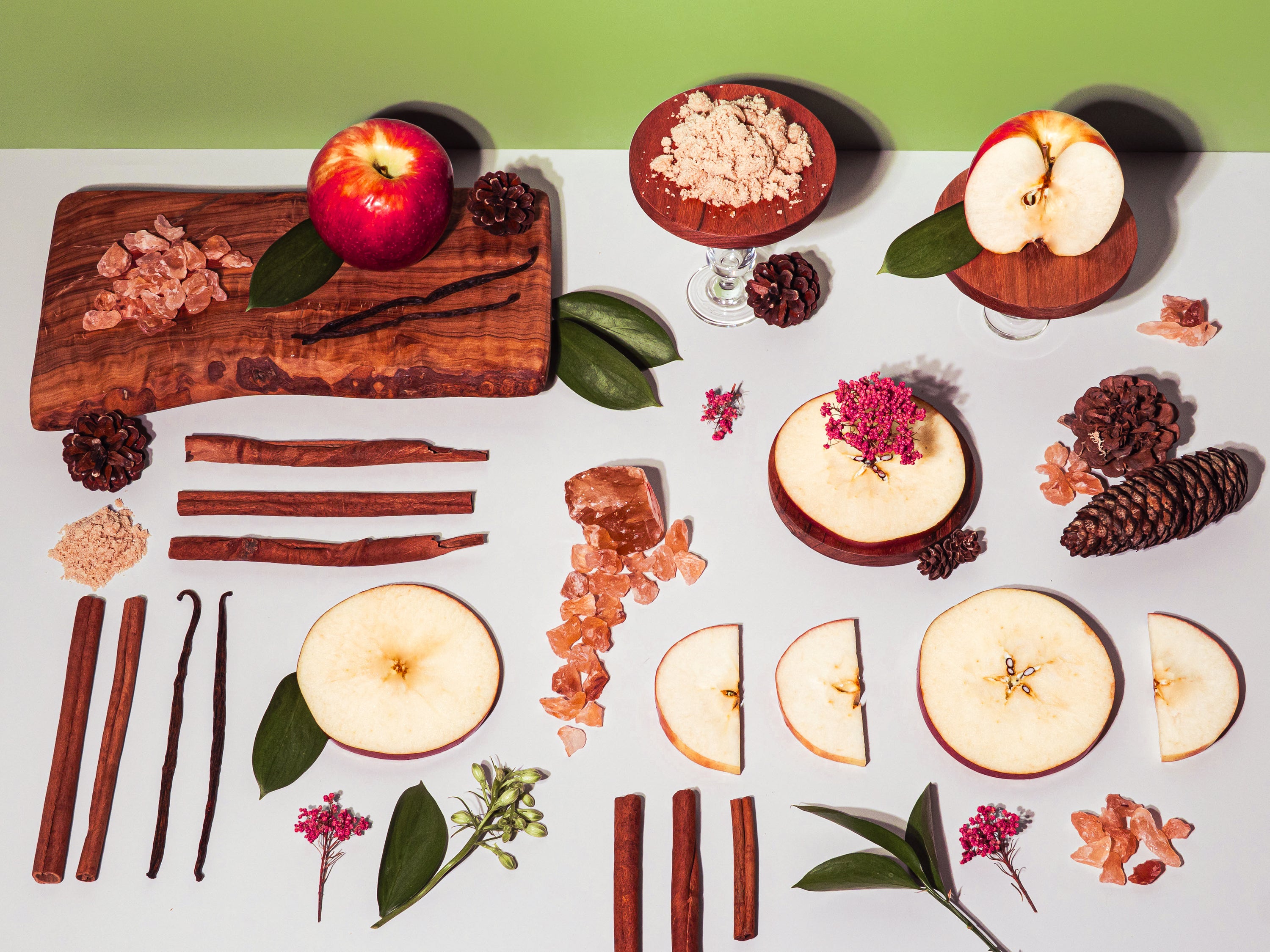Flat lay of fragrance ingredients on a white surface with a green background, featuring whole and sliced apples, cinnamon sticks, vanilla beans, brown sugar, rock sugar crystals, pinecones, and small flowers, arranged in neat rows alongside wooden boards and leaves