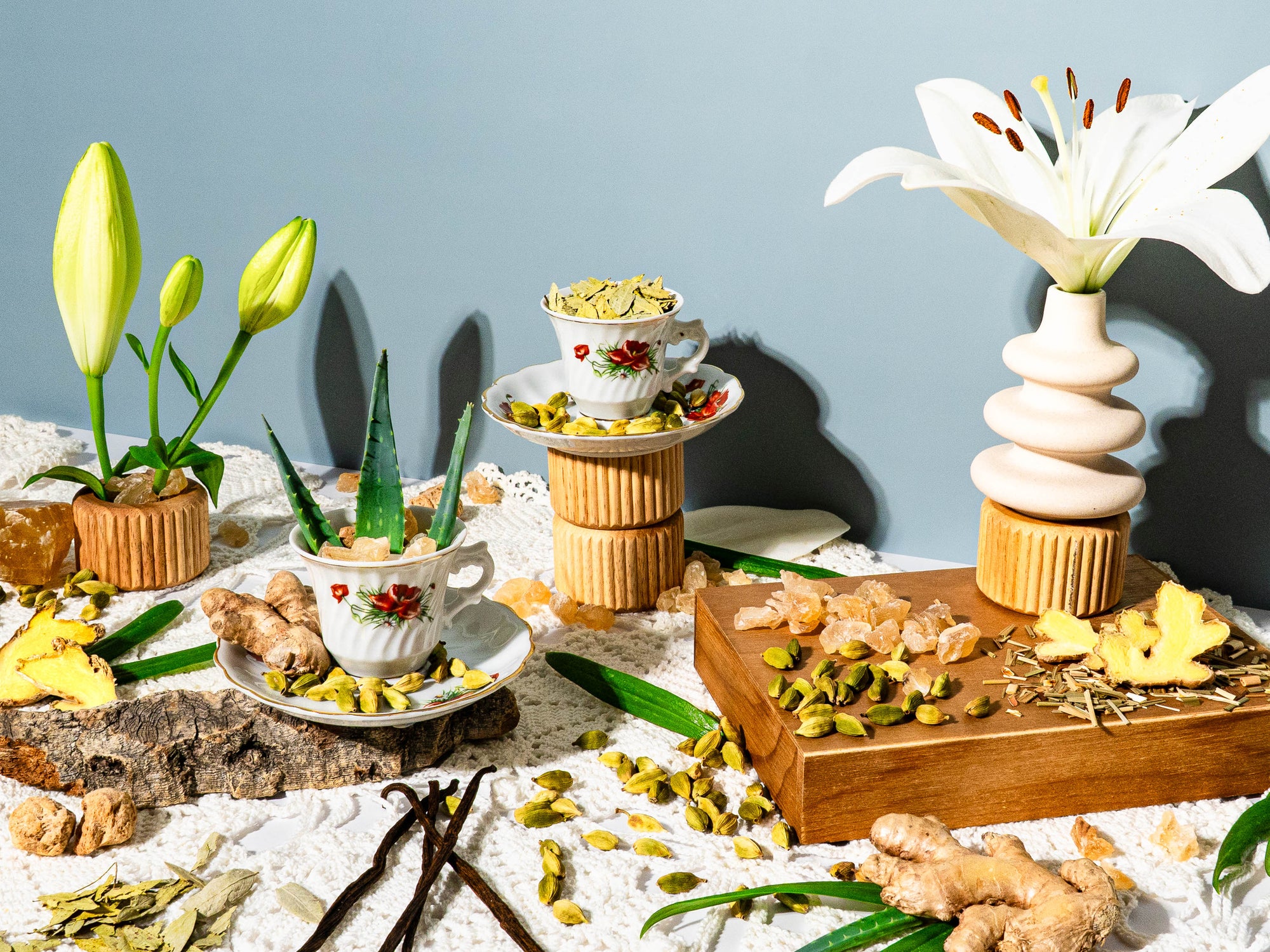 Fragrance oil flat lay featuring ginger root, cardamom pods, aloe vera leaves, vanilla beans, lemongrass, and rock sugar styled with vintage floral teacups and blooming white lilies on a lace-covered table with soft natural lighting.