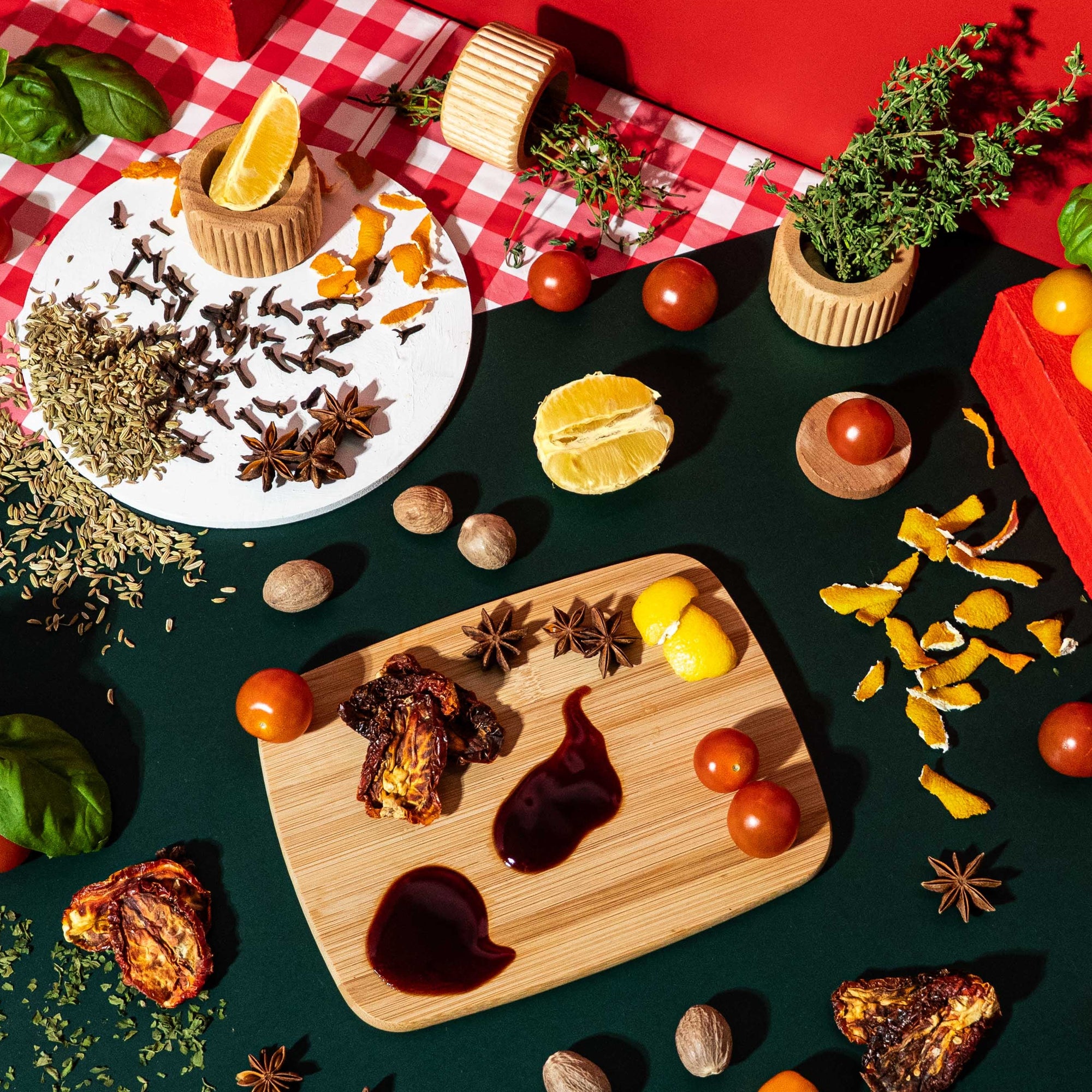 Colorful Italian kitchen inspired flat lay for fragrance oil, featuring fresh basil, cherry tomatoes, thyme, orange and lemon peels, dried herbs, fennel seeds, cloves, star anise, nutmeg, sundried tomatoes, and balsamic vinegar on a dark green surface with red checkered accents and a wooden cutting board.
