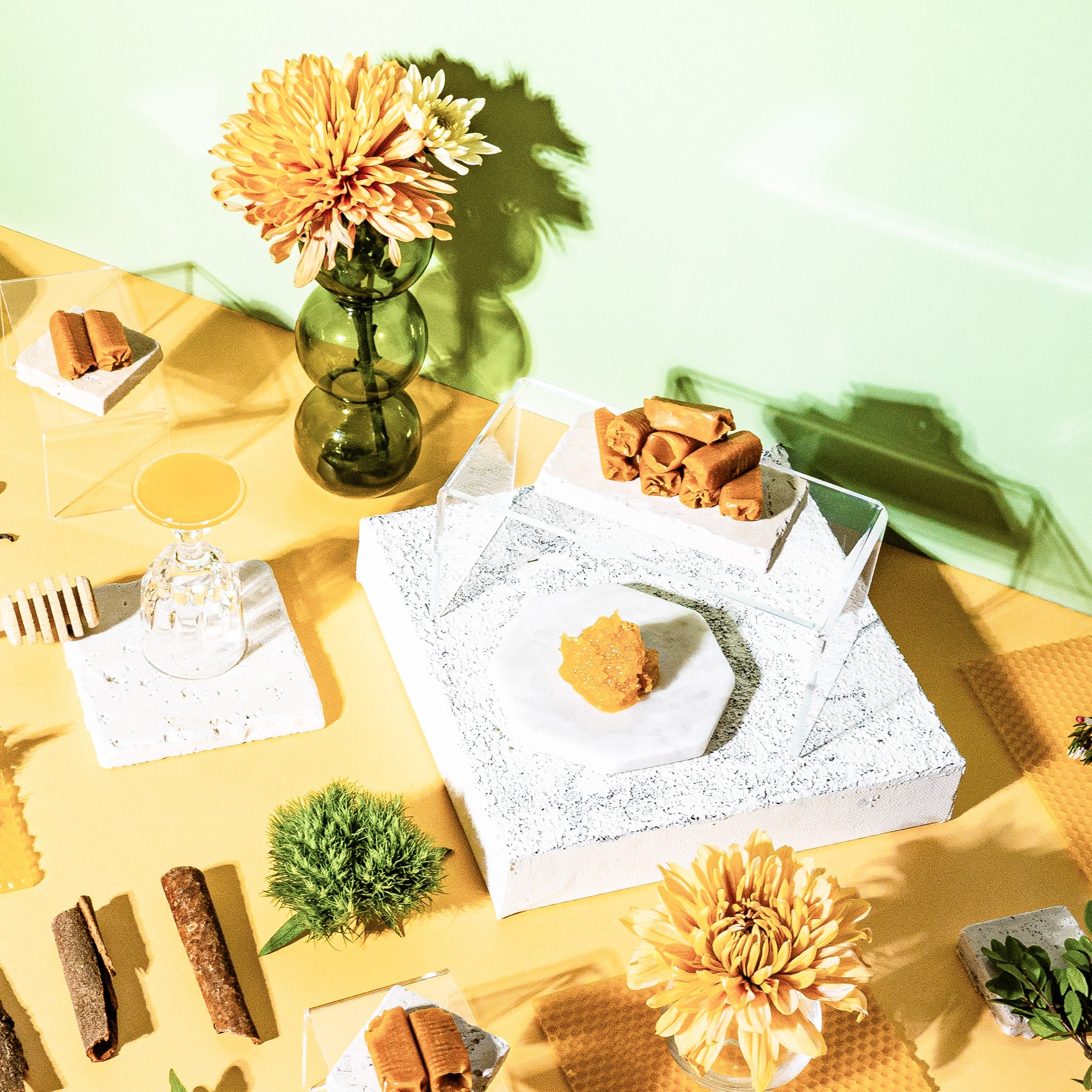 A vibrant flat-lay featuring honeycomb, cinnamon sticks, vanilla beans, and a selection of natural elements like flowers, greenery, and wax materials. The composition evokes warmth and sweetness, with a modern aesthetic showcasing honey drizzler, floral accents, and a soft yellow and green backdrop. A perfect representation of nature-inspired fragrances and cozy indulgence.