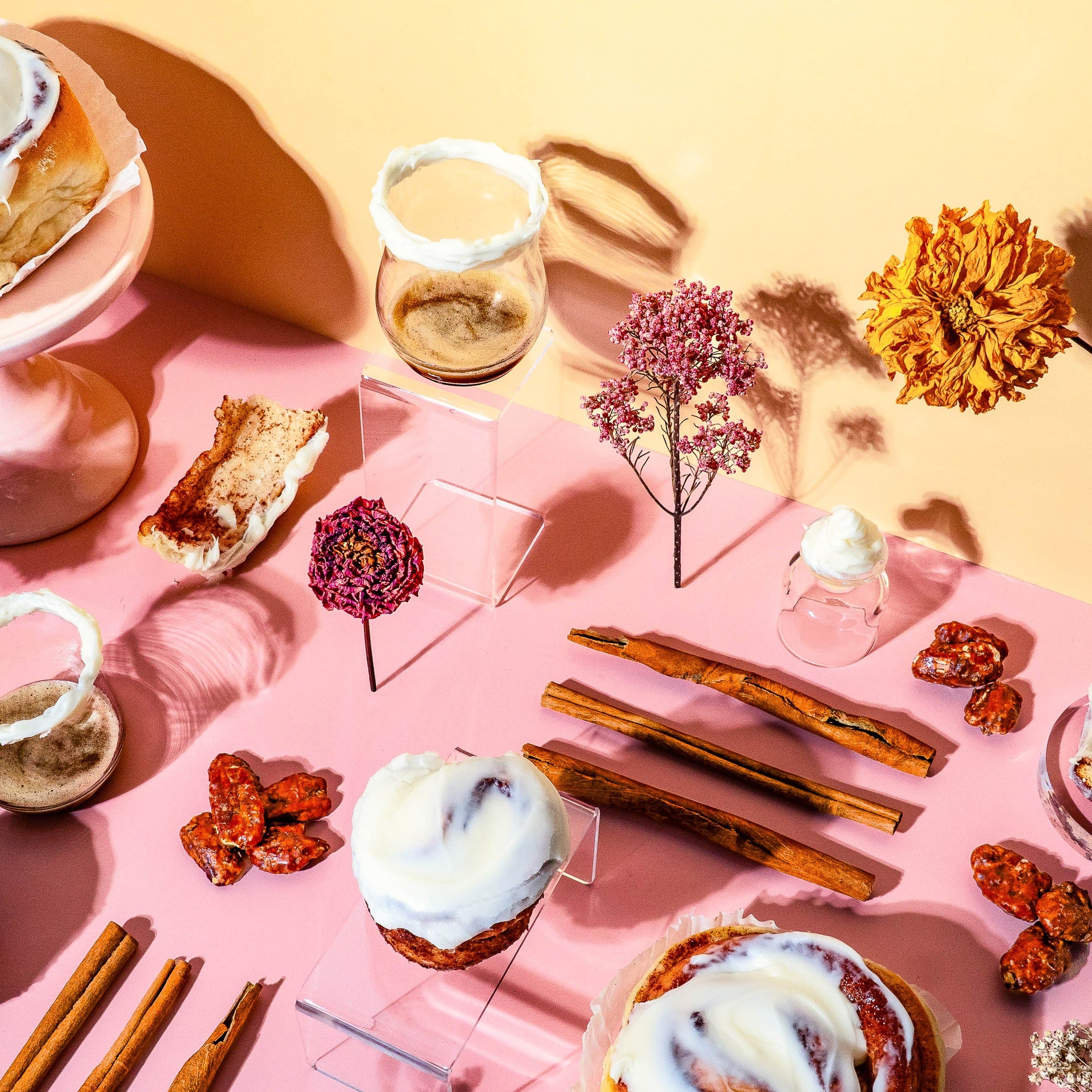 Aesthetic flat lay for a fragrance oil, featuring frosted cinnamon rolls, whole cinnamon sticks, whipped vanilla cream, dried flowers, caramelized nuts, and dessert crumbs arranged on a pink and peach background with modern glass accents and soft natural lighting.