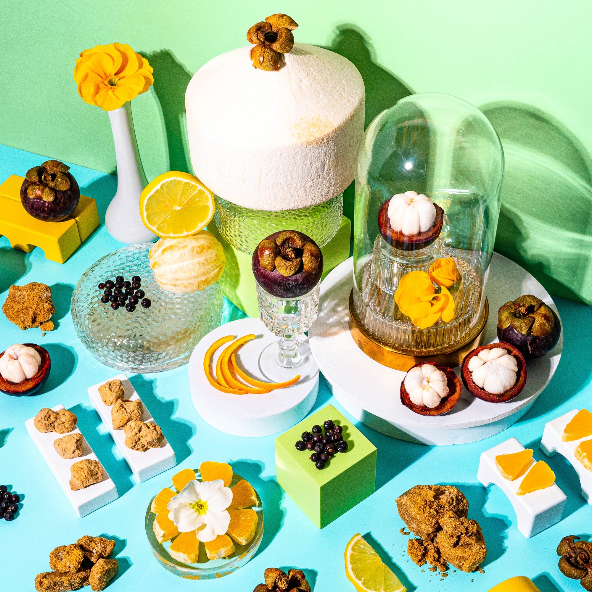 A vibrant flat-lay composition featuring tropical ingredients against a bright turquoise and green background. A whole coconut sits on a textured glass pedestal, surrounded by halved mangosteens, fresh citrus slices, black currants, and golden sugar-coated treats. A glass cloche encases a single mangosteen and delicate yellow flowers, adding an elegant touch. Scattered lemon slices, orange peels, and white blossoms enhance the summery, sunlit feel of the scene, evoking the a fresh, tropical fragrance.