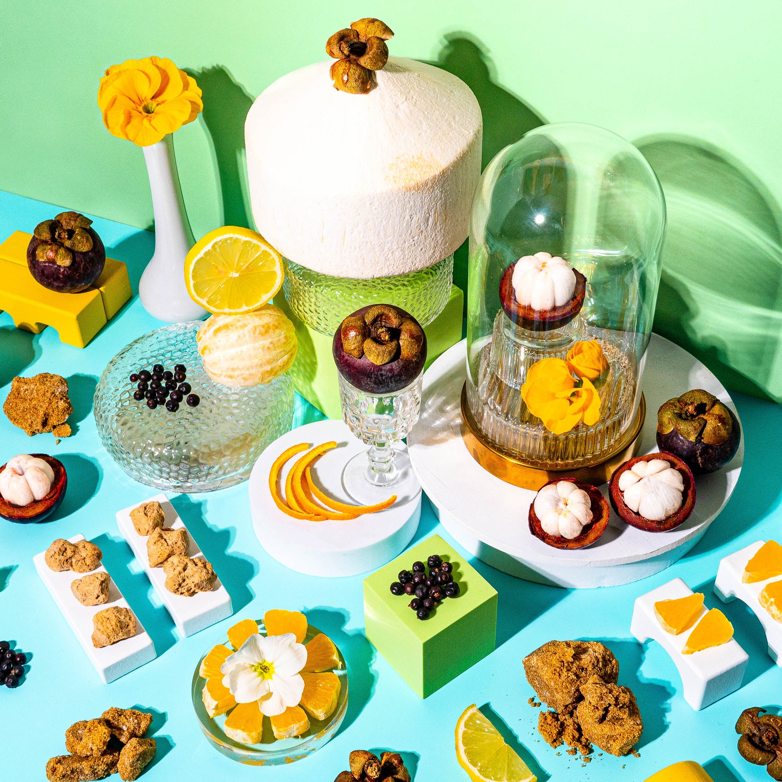 A vibrant flat-lay composition featuring tropical ingredients against a bright turquoise and green background. A whole coconut sits on a textured glass pedestal, surrounded by halved mangosteens, fresh citrus slices, black currants, and golden sugar-coated treats. A glass cloche encases a single mangosteen and delicate yellow flowers, adding an elegant touch. Scattered lemon slices, orange peels, and white blossoms enhance the summery, sunlit feel of the scene, evoking the a fresh, tropical fragrance.