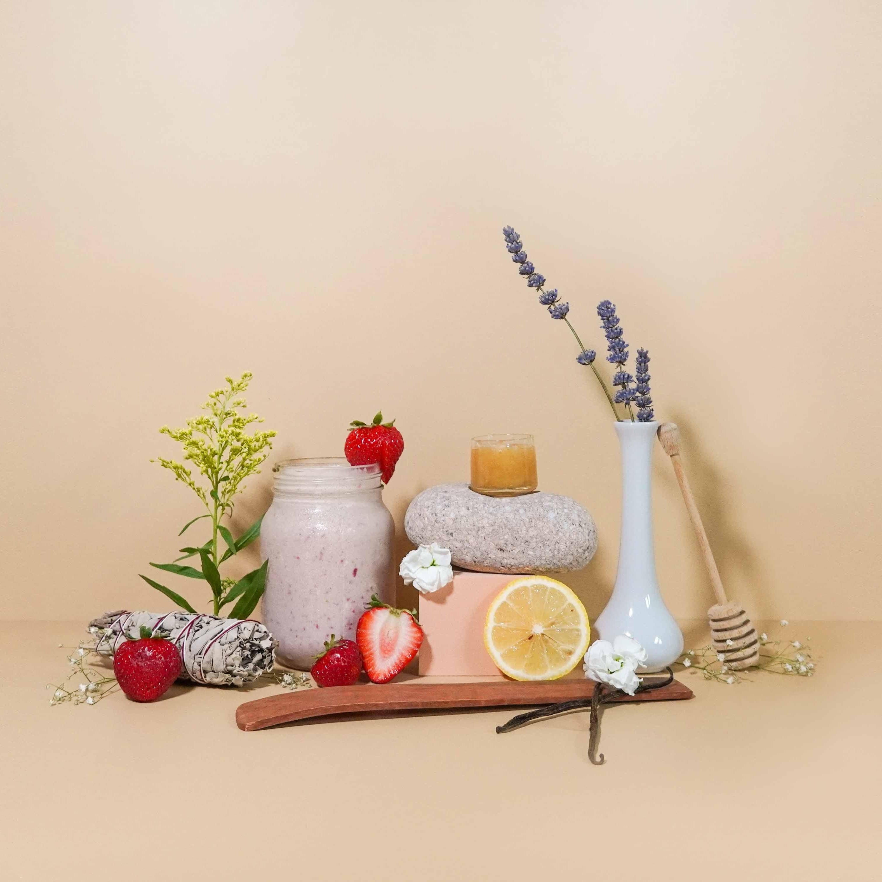 An arrangement of fragrance oil bottles, a lemon, strawberries, a lemon slice, a strawberry slice, flowers, and greenery against a neutral background.