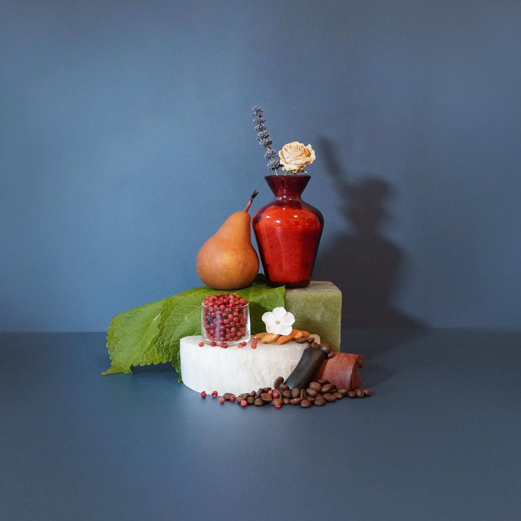 A fragrance oil display with a red bottle, surrounded by a slice of pear, a piece of bread, and coffee beans, presented on a blue background.