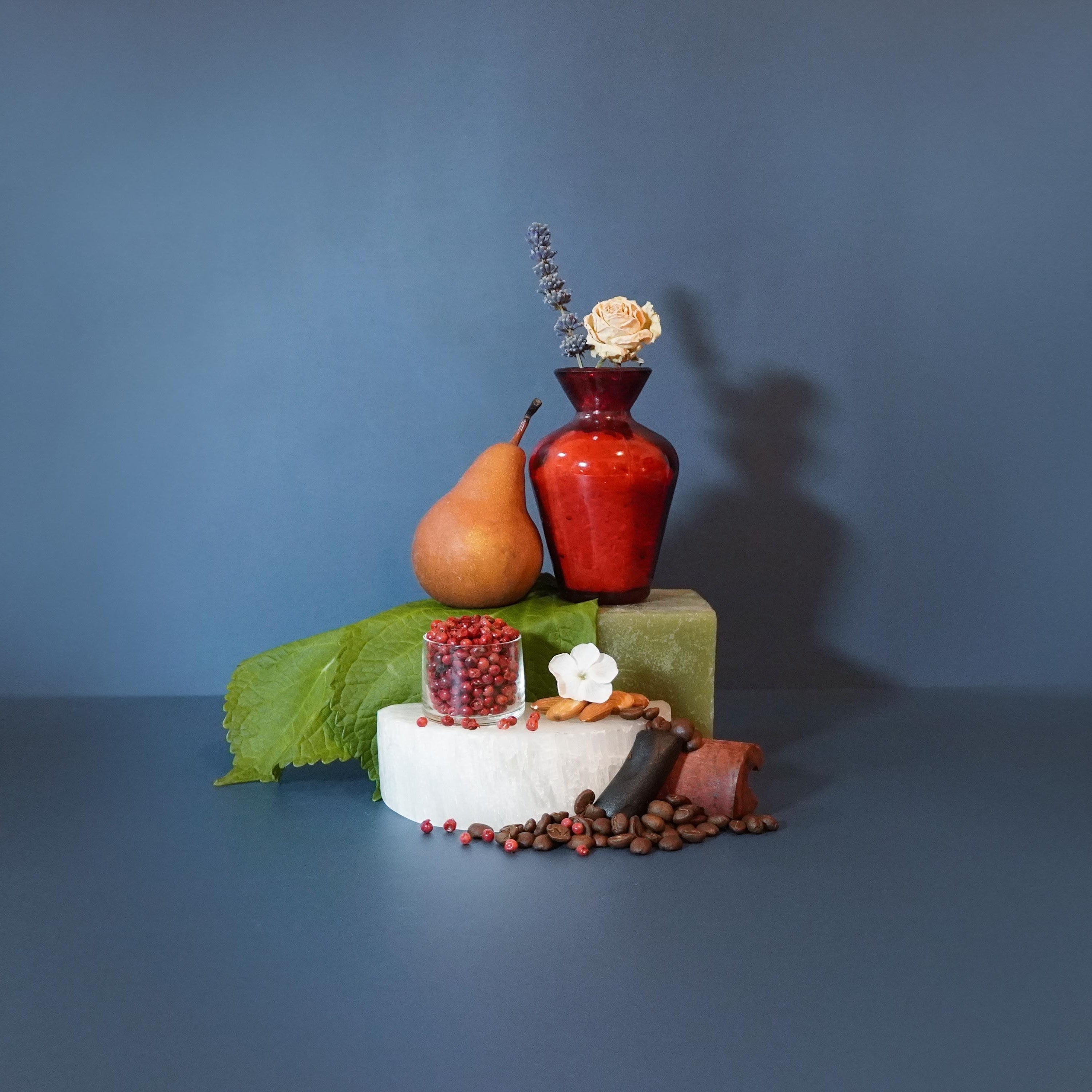 A fragrance oil display with a red bottle, surrounded by a slice of pear, a piece of bread, and coffee beans, presented on a blue background.