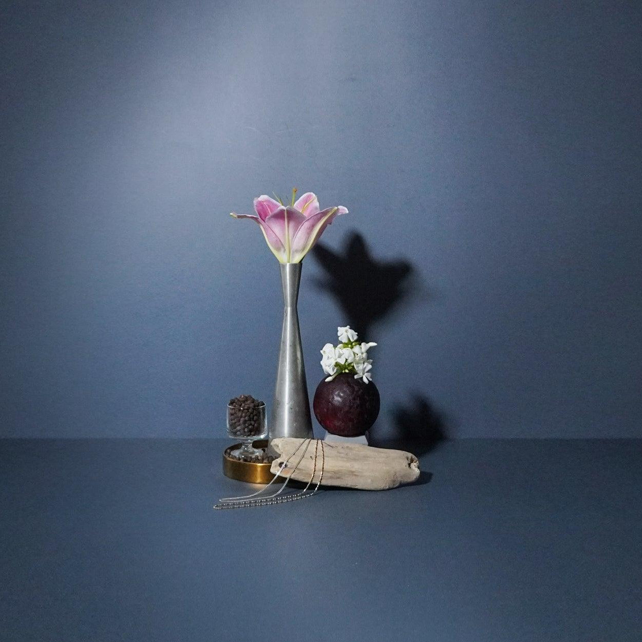 A metal candle sick, alongside a small glass container, displayed on a dark background with a decorative flower and a shadow cast on the wall.