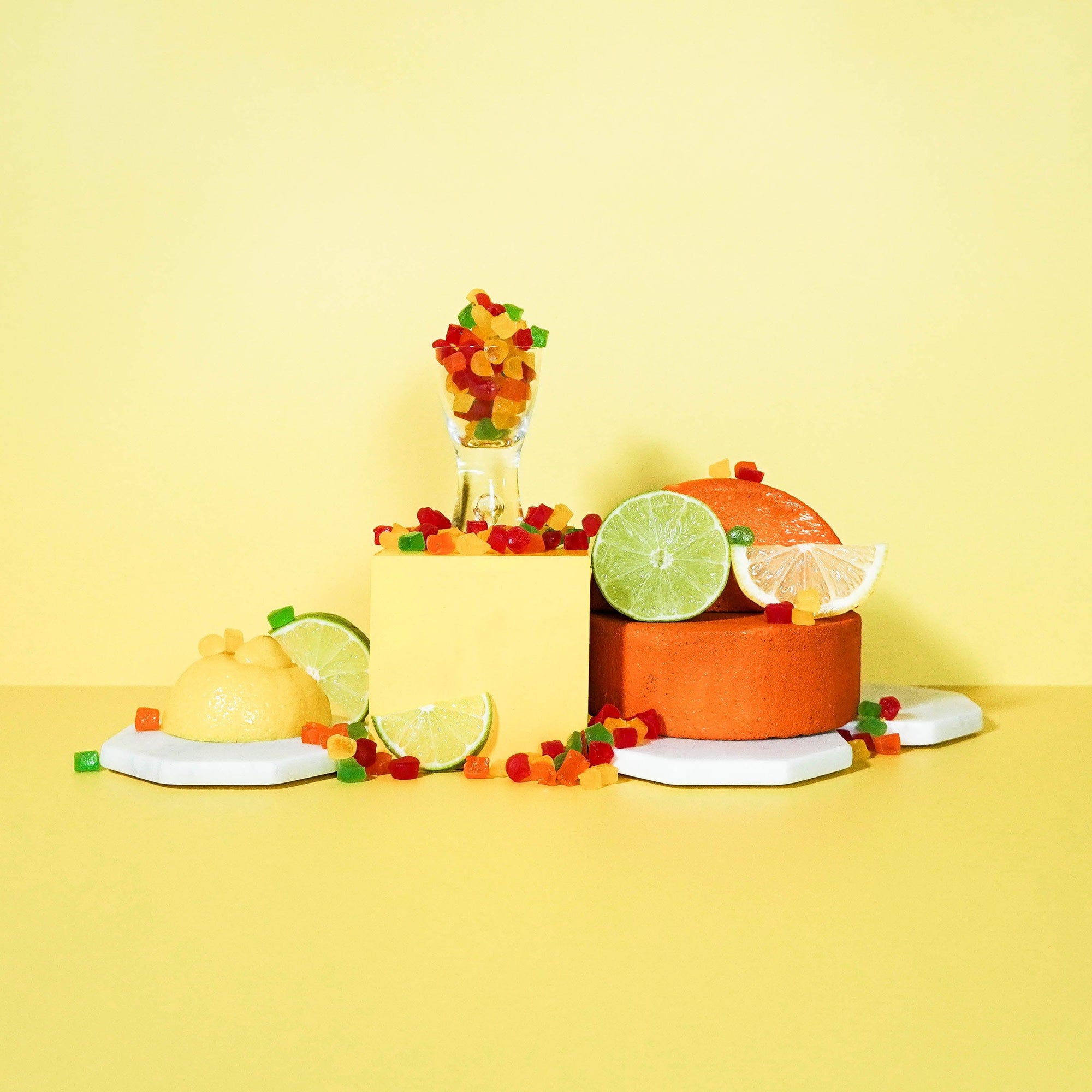 A still life setup featuring a small clear bottle with a red cap, surrounded by citrus fruits like lemon and lime, and decorative elements like raspberries and a flower, all arranged on a yellow background.