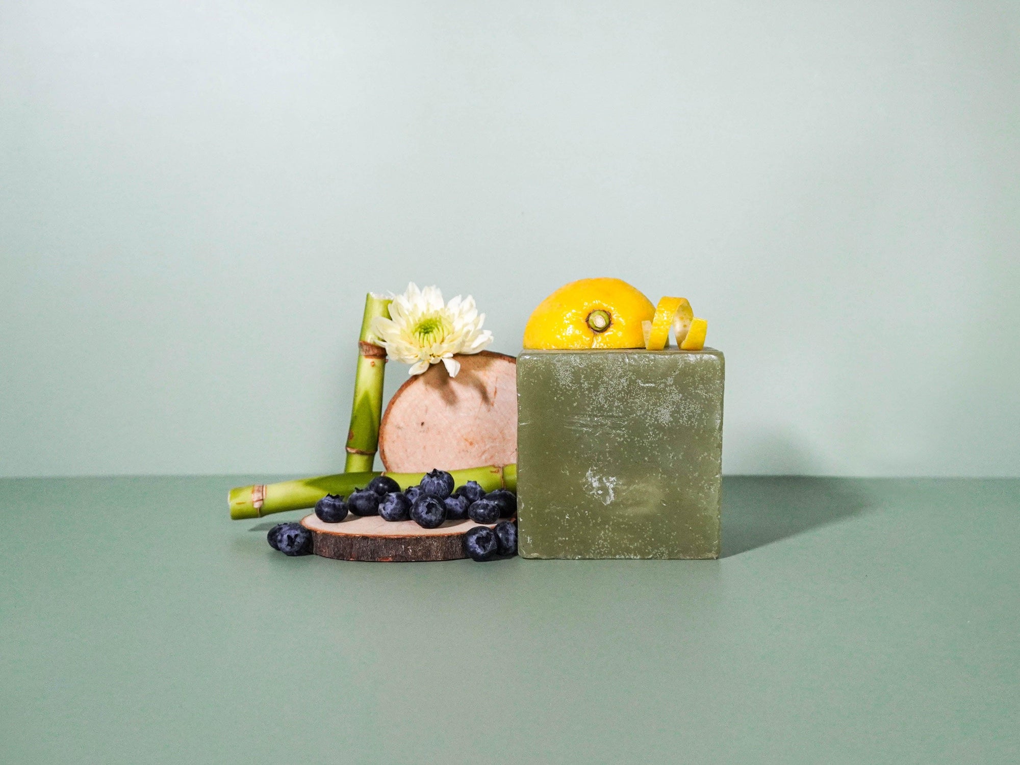 A bar of soap with a greenish hue and decorative elements including a lemon, a huckleberry, and a vanilla pod arranged around it on a green background.