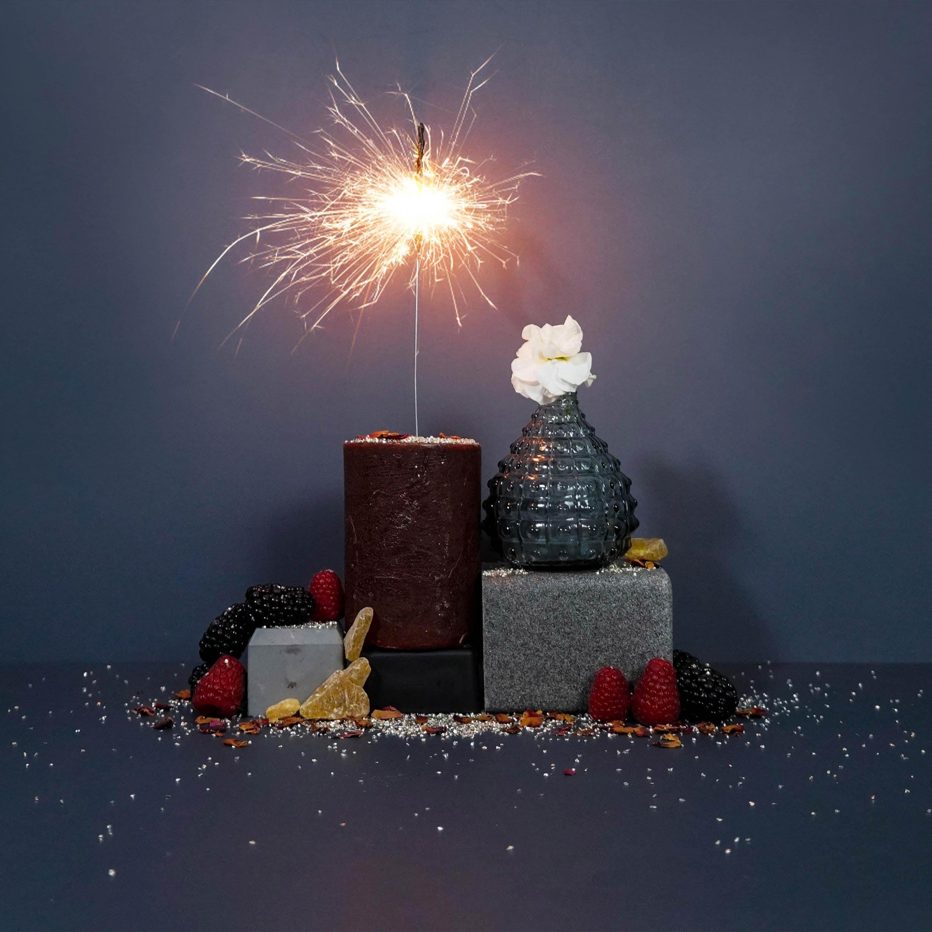 A fragrance oil bottle with a sparkler, surrounded by decorative berries and a flower on a dark background.