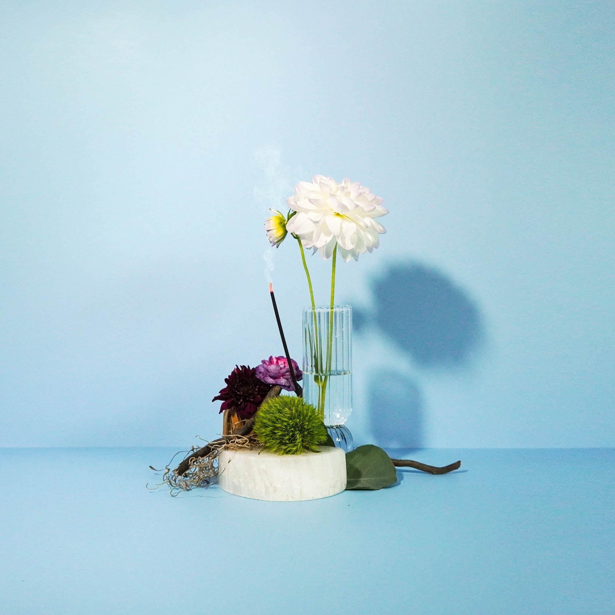 A small glass vase with a bouquet of flowers, placed on a white surface with a blue background. The flowers include a white dahlia, pink and purple flowers, and green foliage.