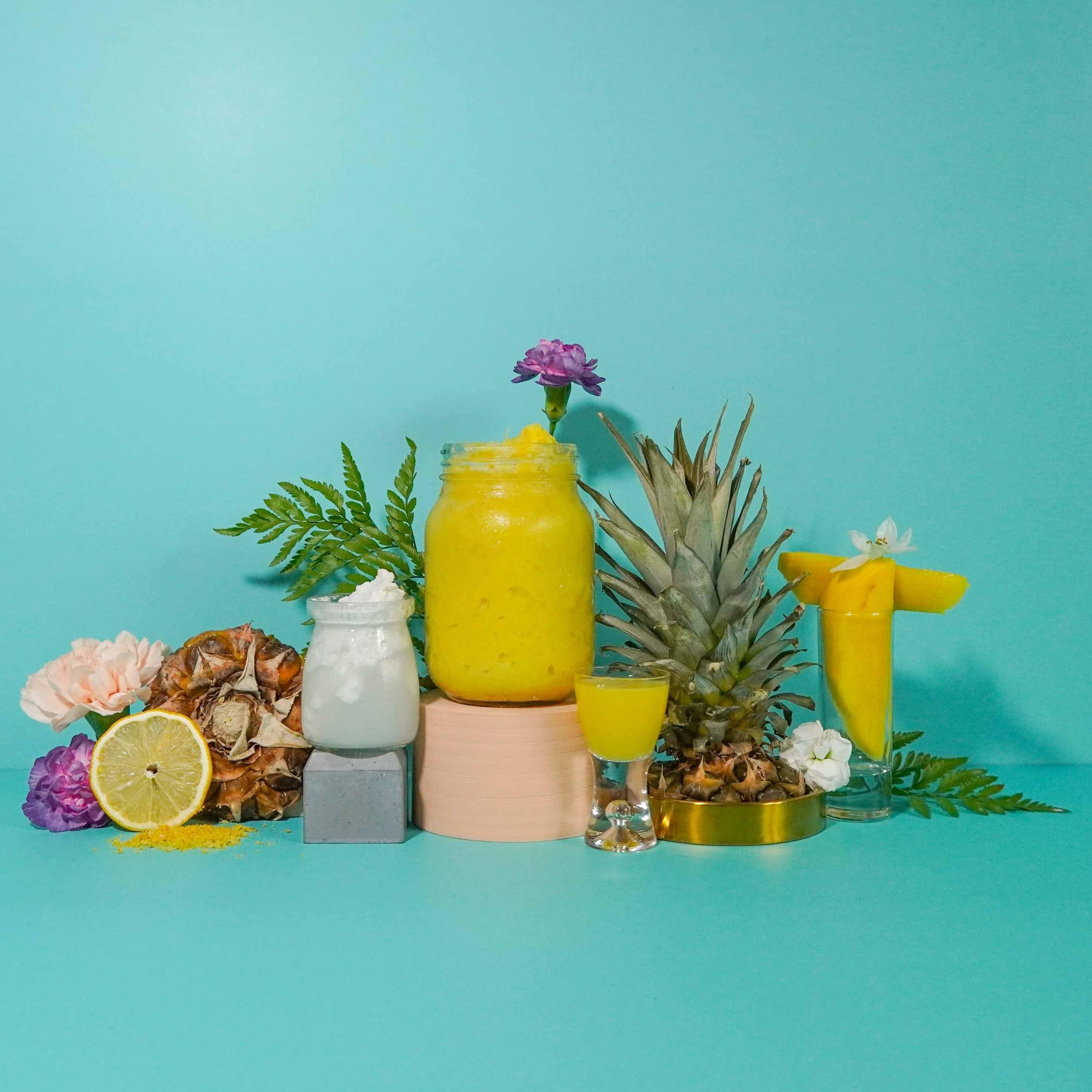 A display of various containers with a yellow liquid, surrounded by props including a pineapple, flowers, and citrus fruit, set against a turquoise background.